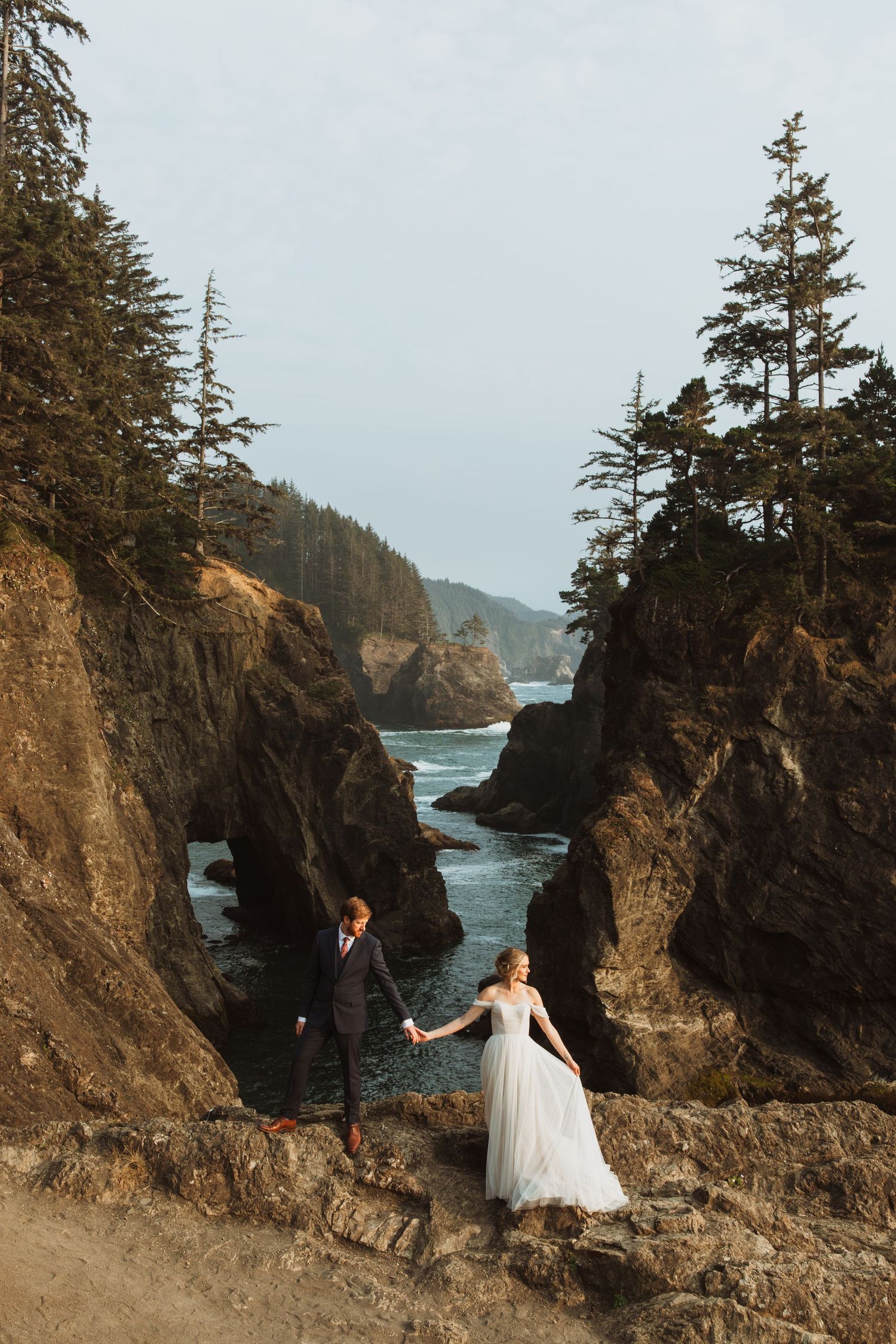 A wedding couple stands on dramatic coastal cliffs with natural rock arches and evergreen trees along the Pacific coast.