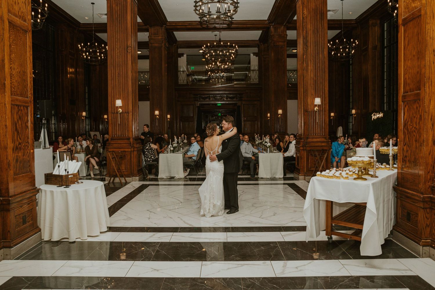 A newlywed couple dancing in an elegant ballroom with marble floors and wooden columns.