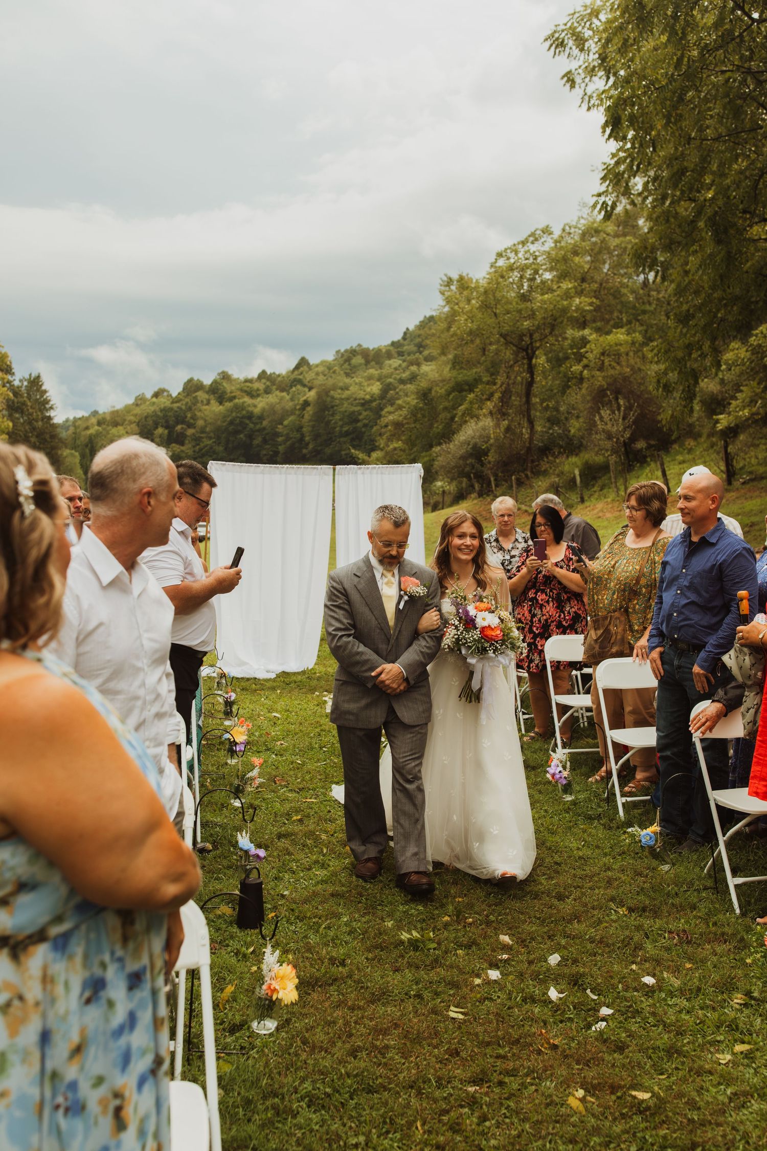 Outdoor wedding ceremony with guests gathered on a grassy hillside under cloudy skies.