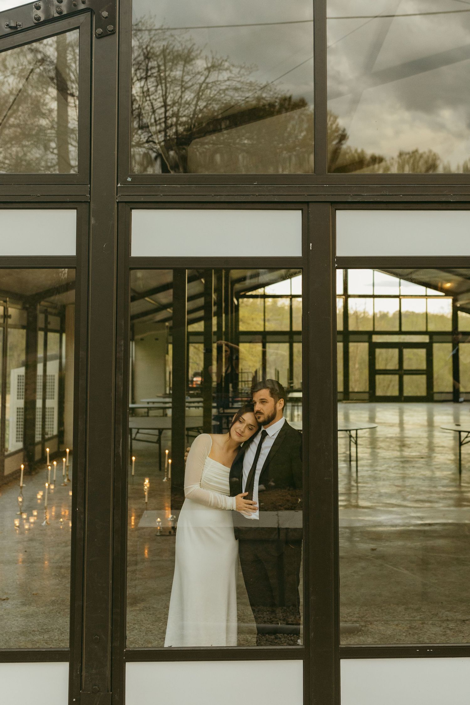 A wedding couple embraces by large windows in an industrial-style venue with string lights.