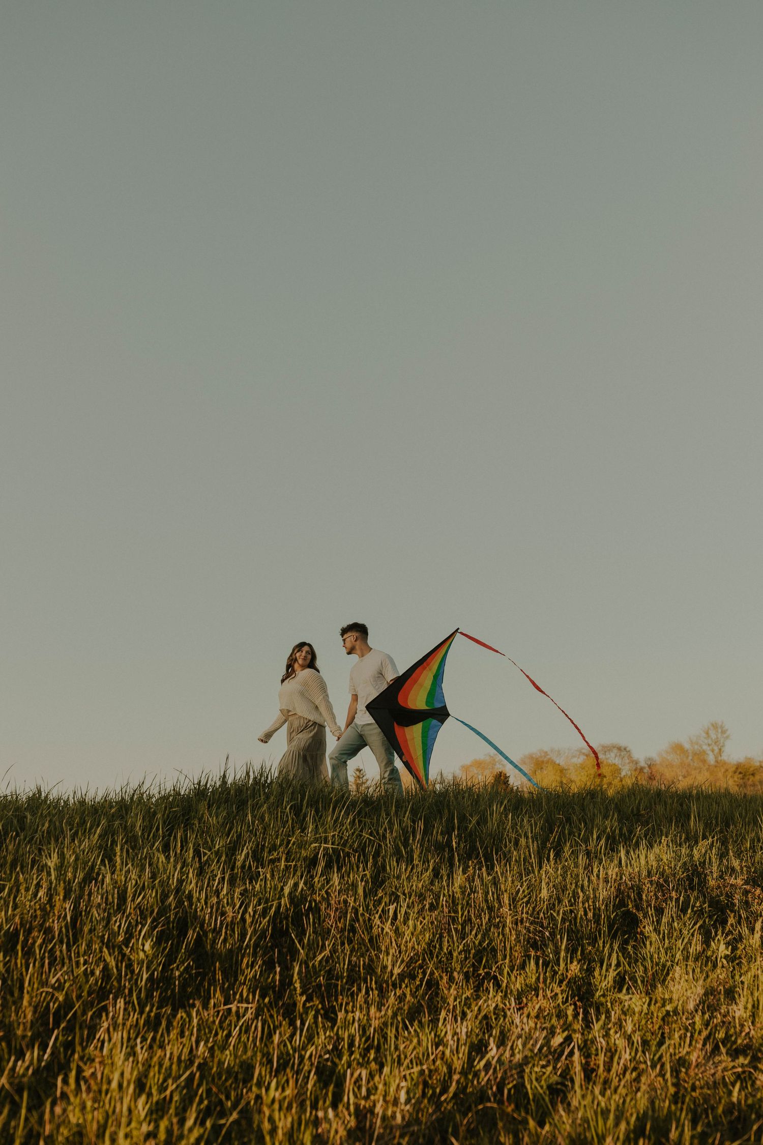 Two figures stand on a grassy hill at sunset with a vibrant rainbow kite.