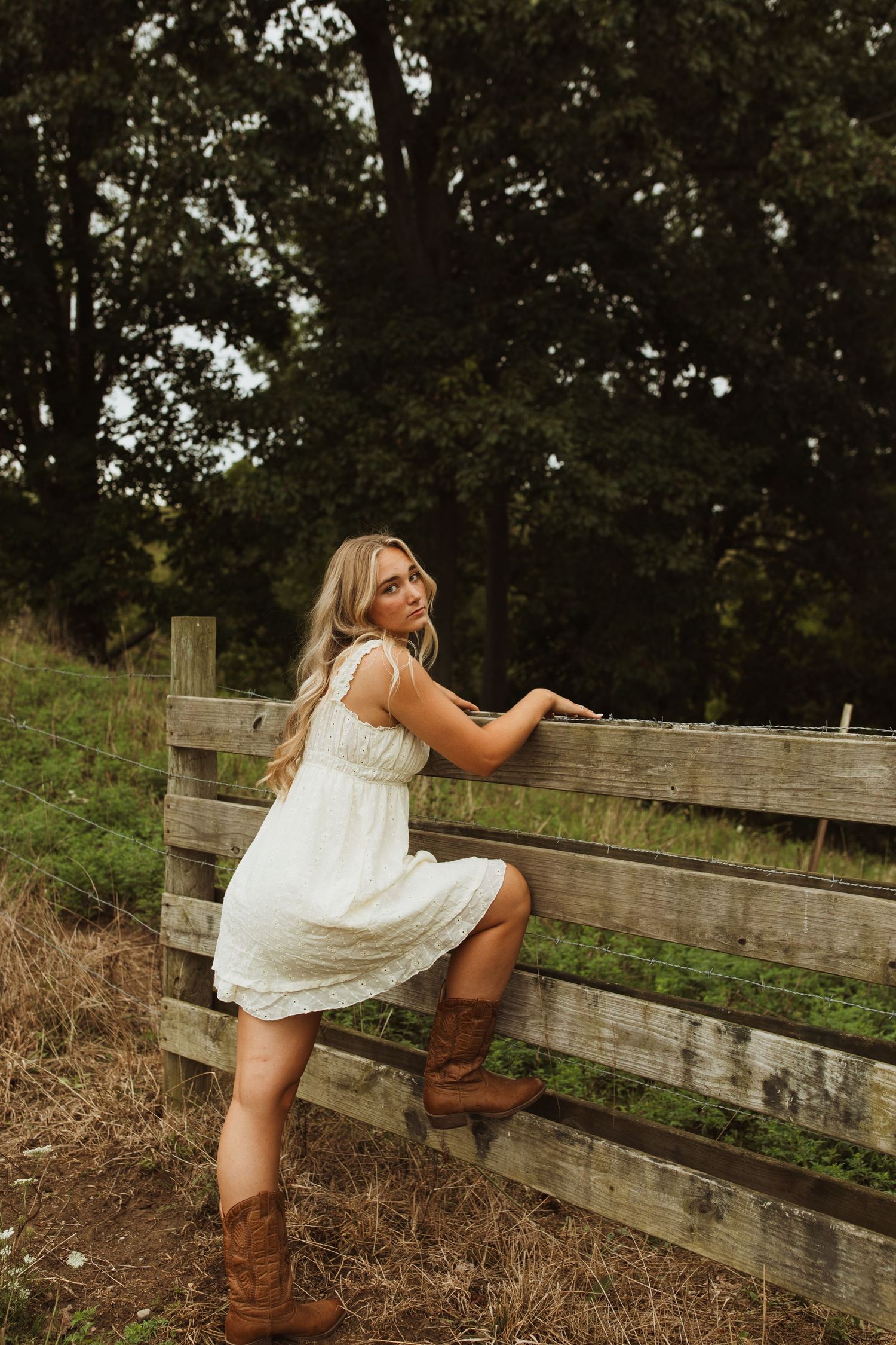 Person in white sundress climbs rustic wooden fence wearing brown boots.
