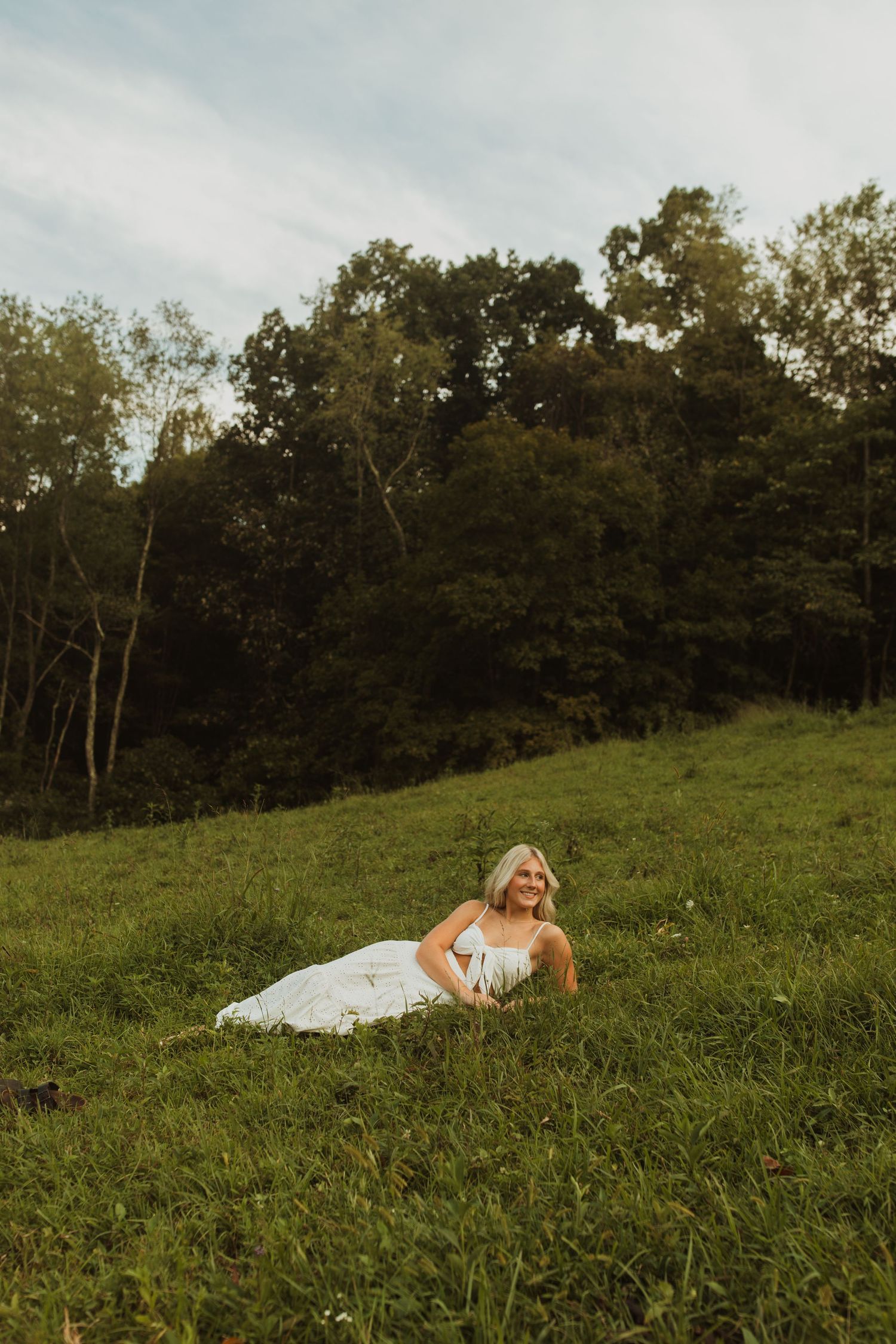 Person in white dress lounges on grassy hillside during sunset.