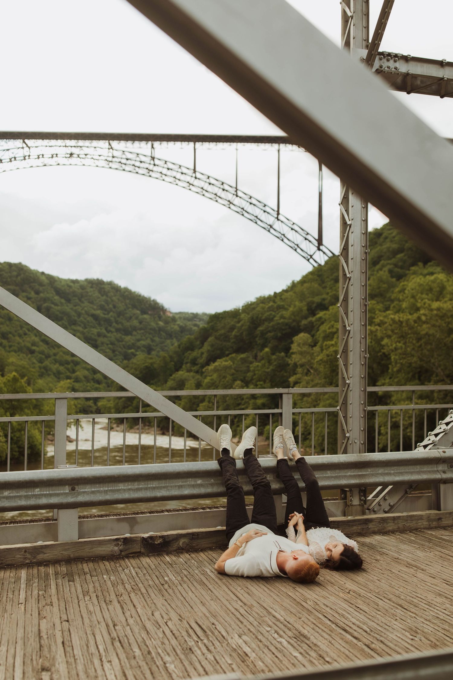 Two people lying the fayette station bridge, with a view of the new river gorge bridge