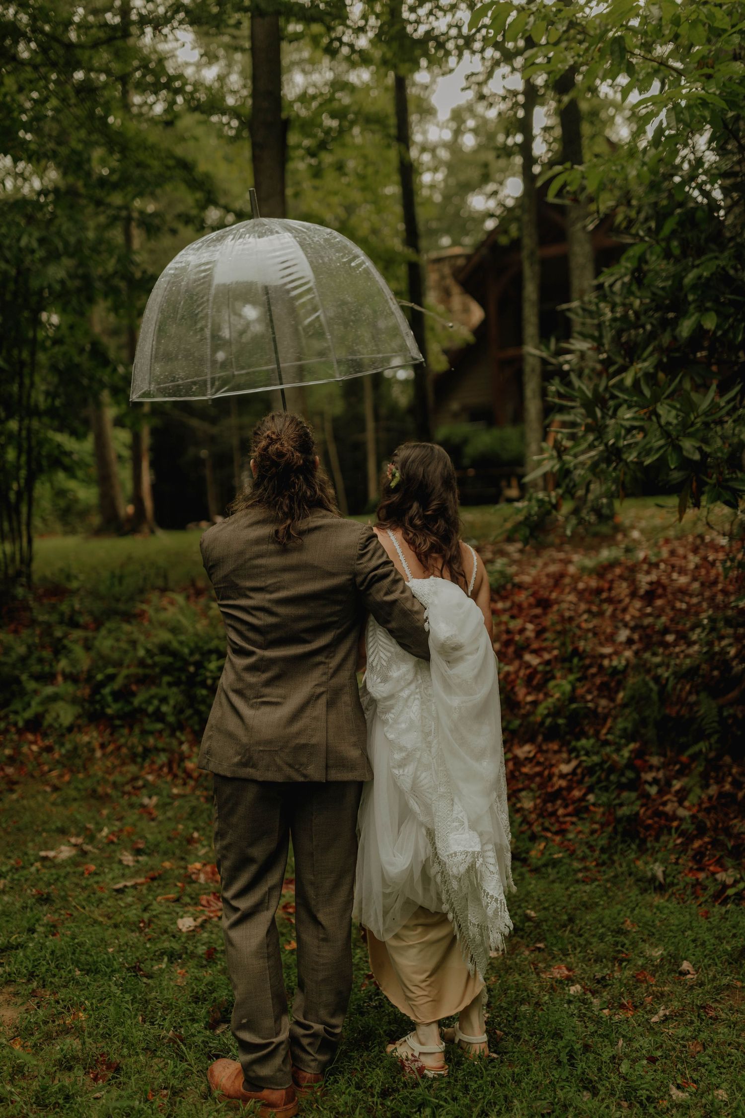 A back view of newlyweds sharing an intimate moment under a clear umbrella in the woods.