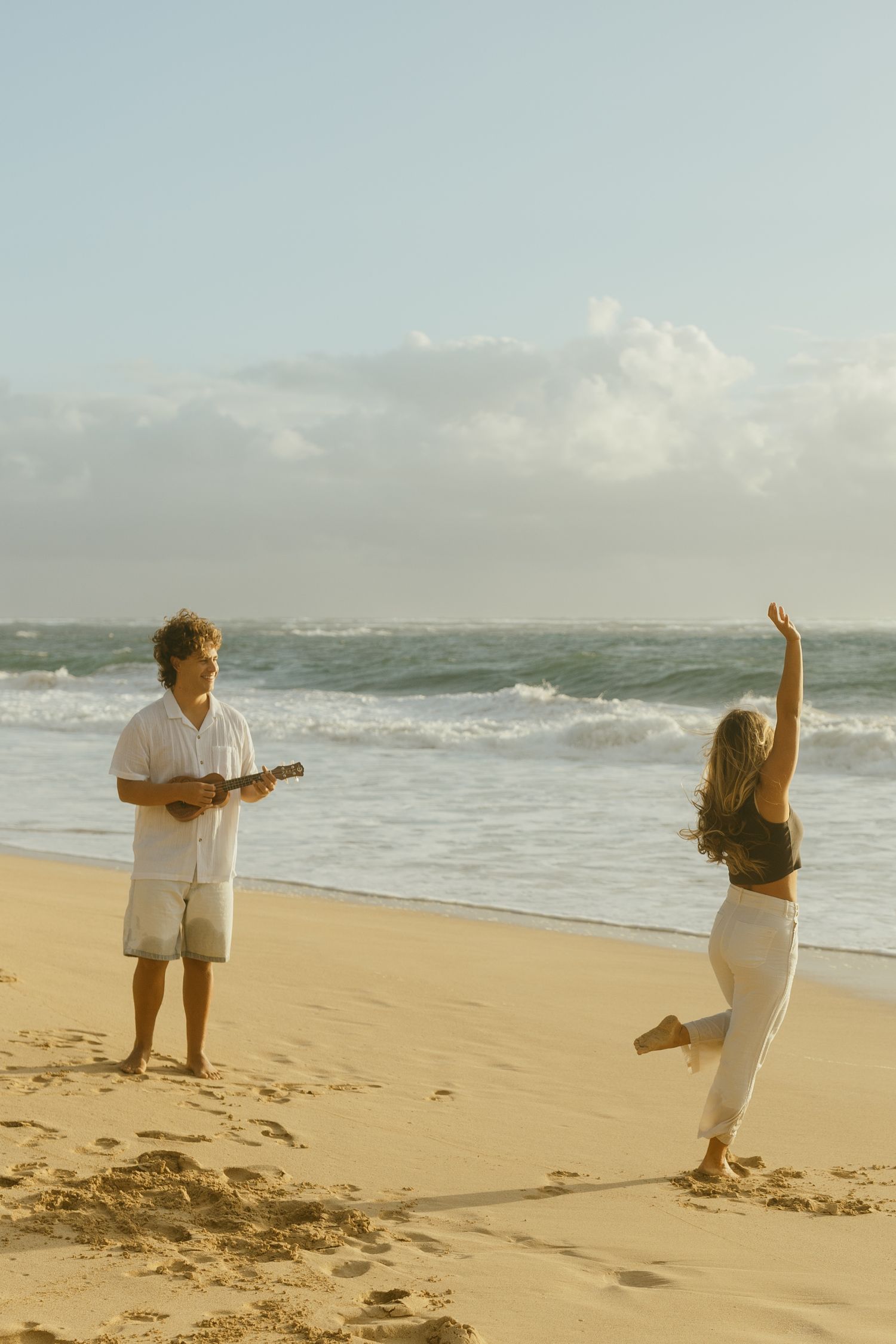 Two people dancing and playing ukulele on a sandy beach at sunset.