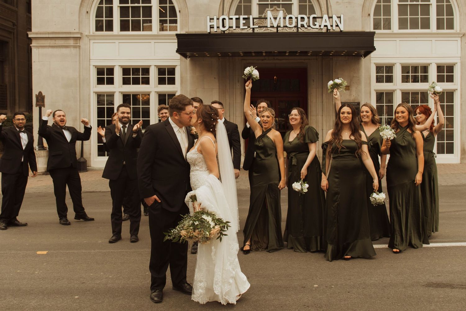 Wedding party poses joyfully in front of Hotel Morgan with bridesmaids wearing elegant dark gowns.