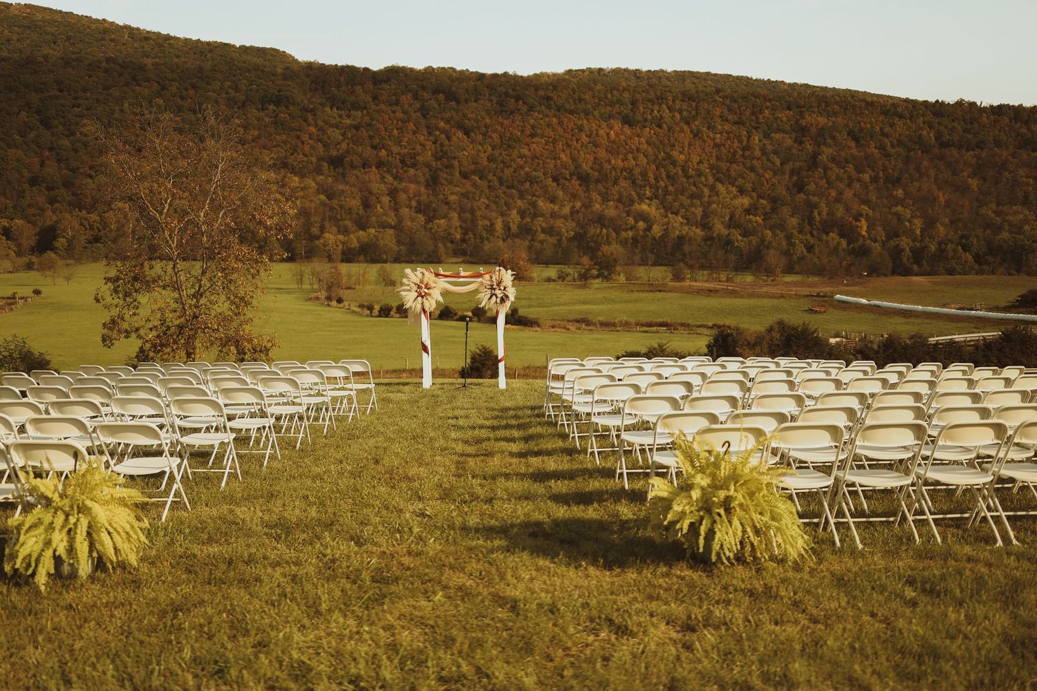 White ceremony chairs arranged in rows leading to a wooden arch on a hillside for an outdoor wedding.