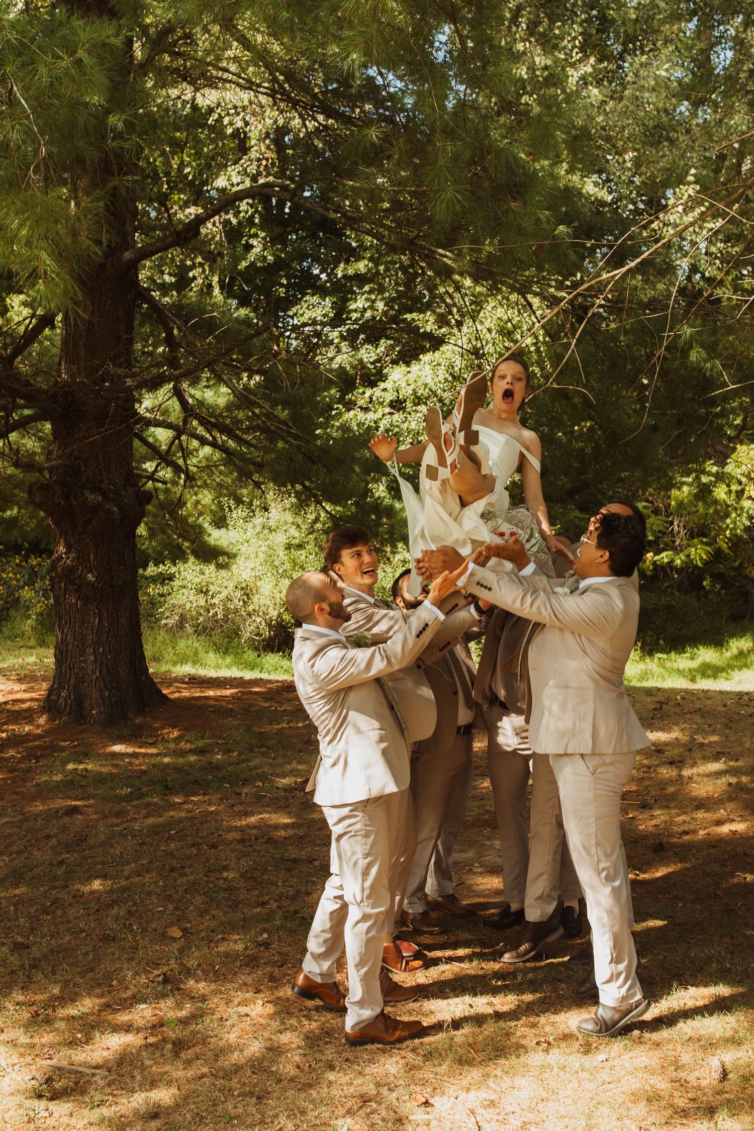 Wedding party members in cream suits lifting someone up in a playful group photo outdoors.