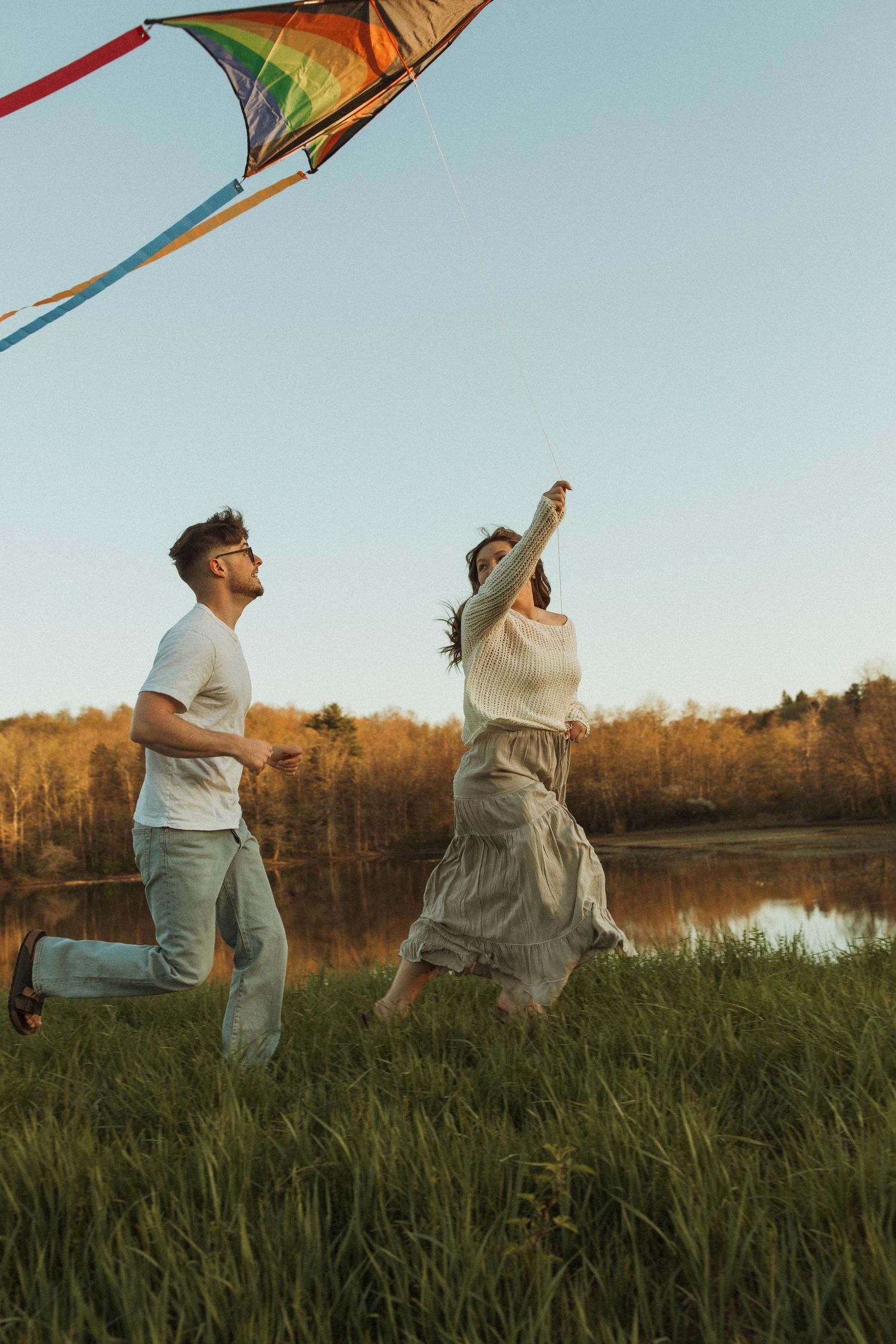 Two people run joyfully through a field while flying a rainbow kite near a lake.