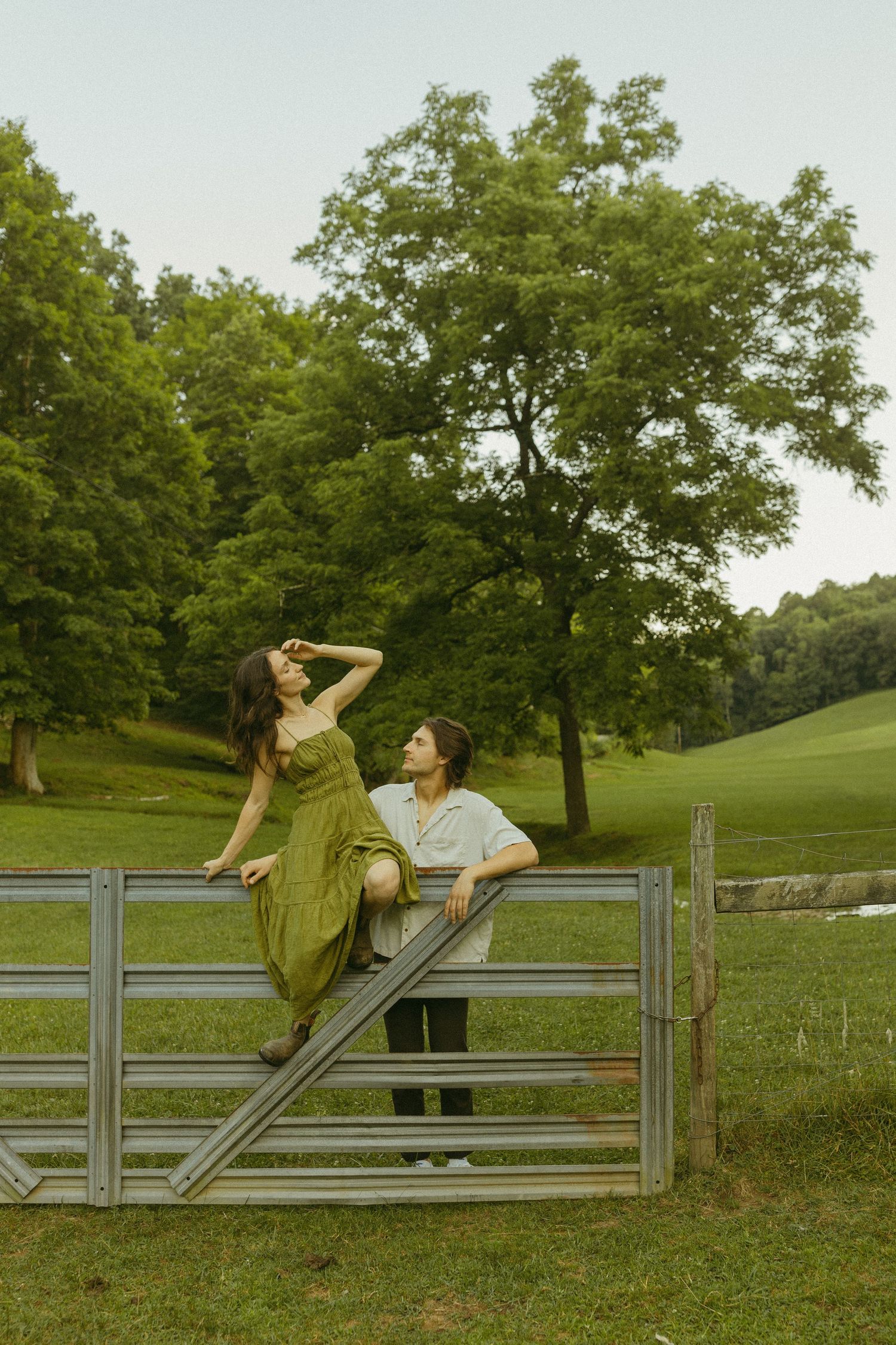 Couple posing playfully on a metal gate in a scenic countryside setting.