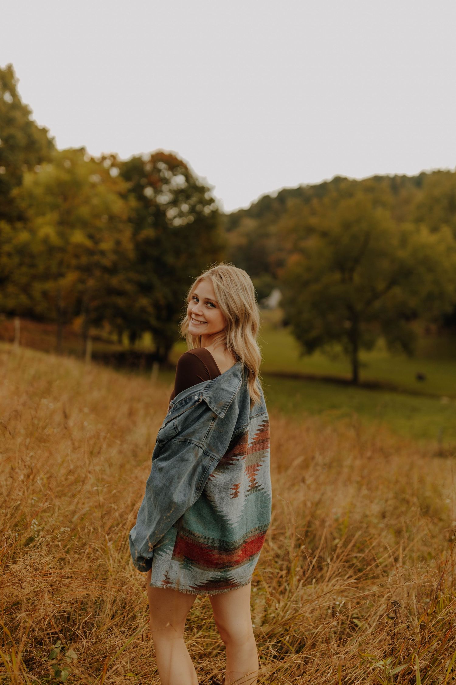 Woman wrapped in patterned blanket stands in golden autumn field at sunset.