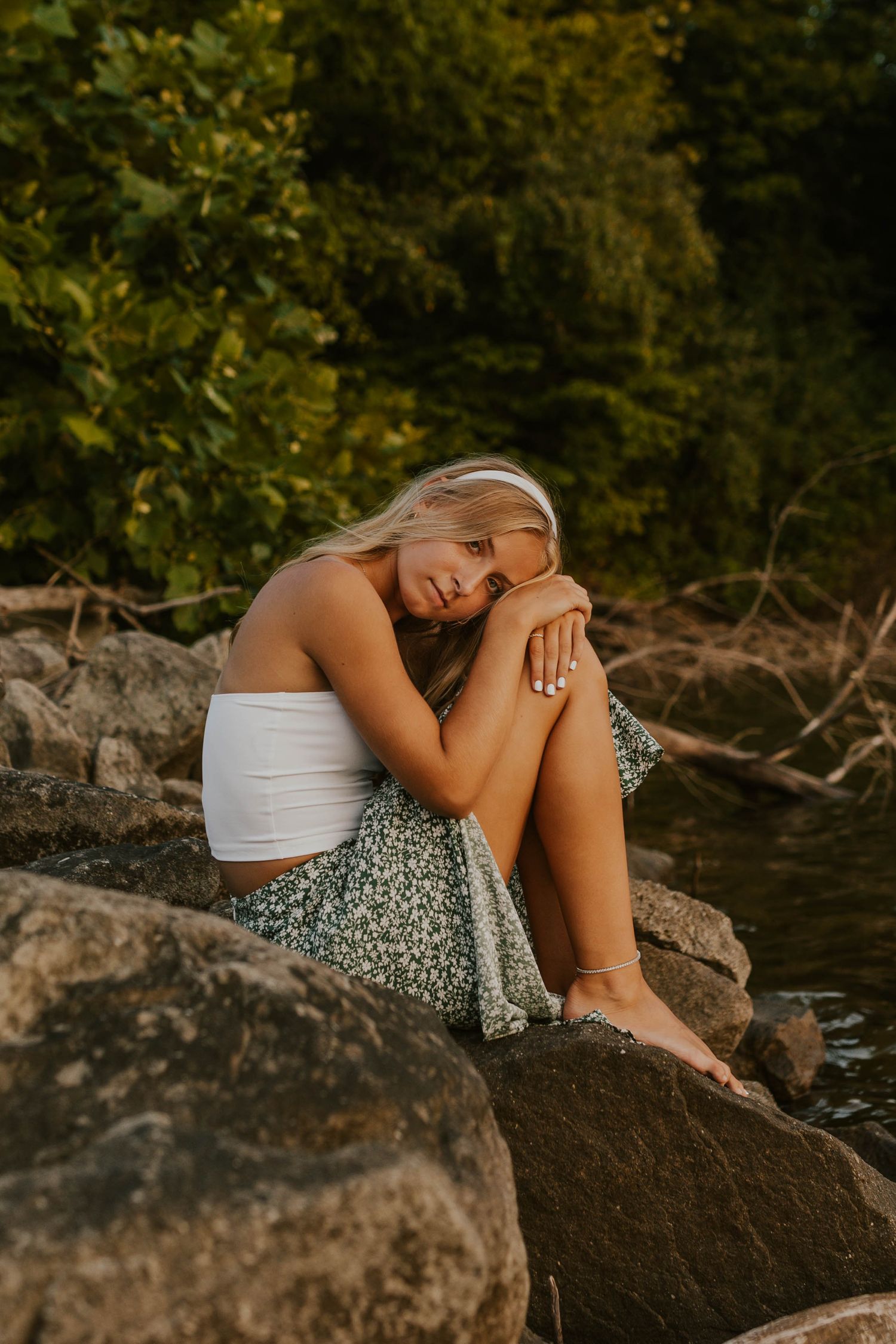 Woman in white top sits contemplatively on rocky shore by lake.