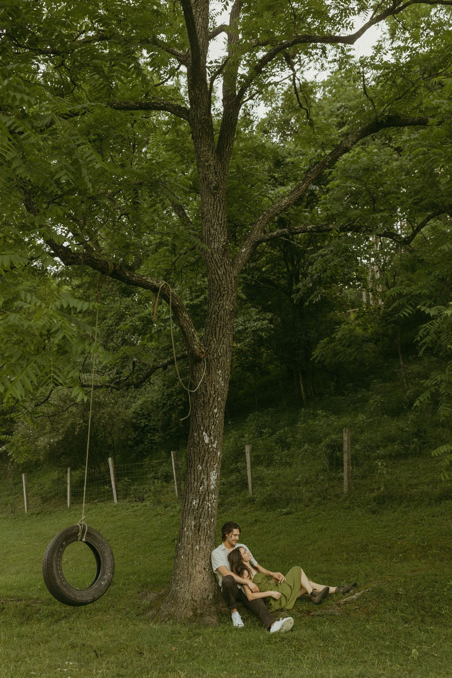 A couple plays on a tire swing in a peaceful park setting during late afternoon.