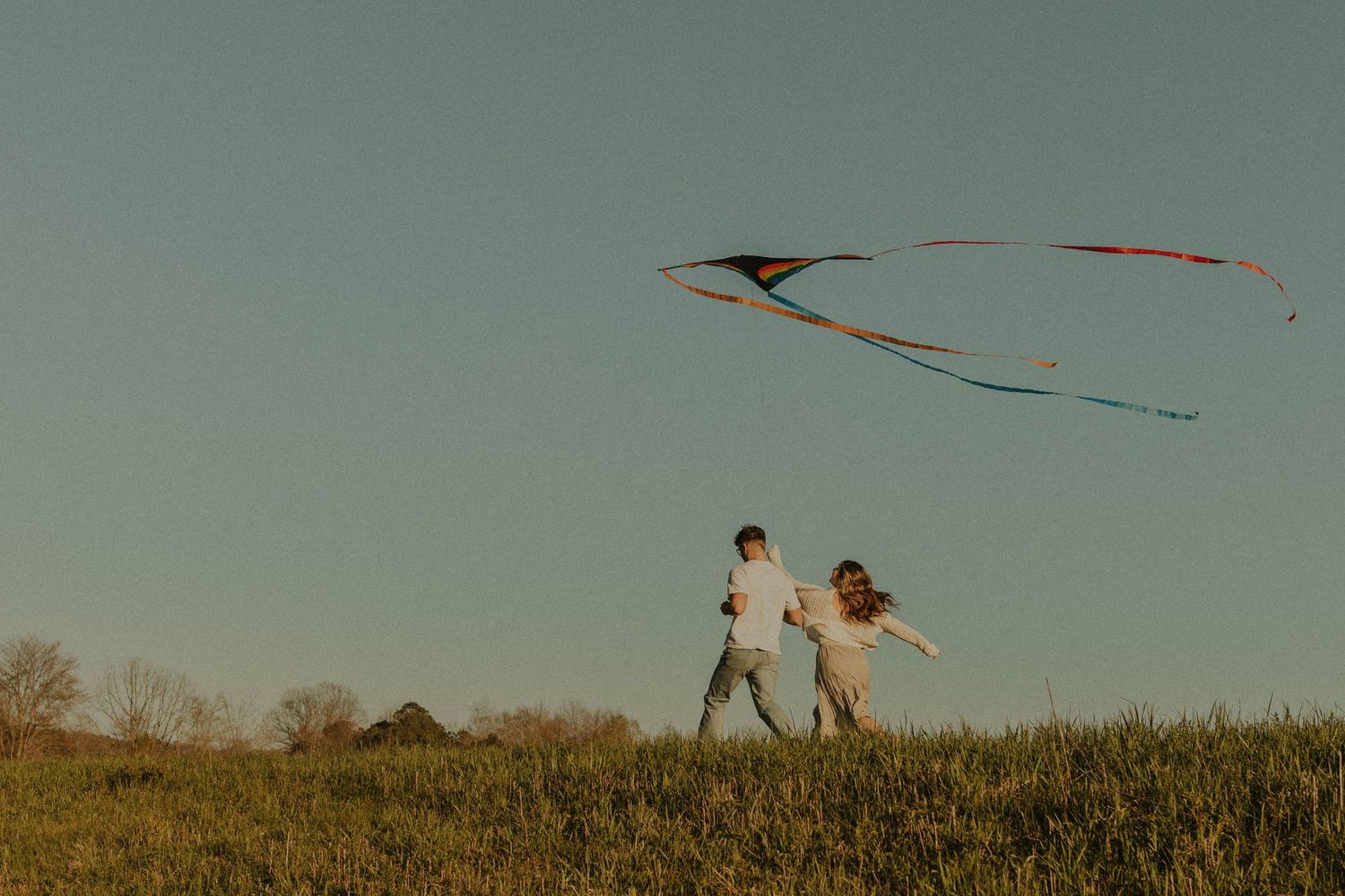 Two people fly a colorful kite together on a hilltop at dusk.