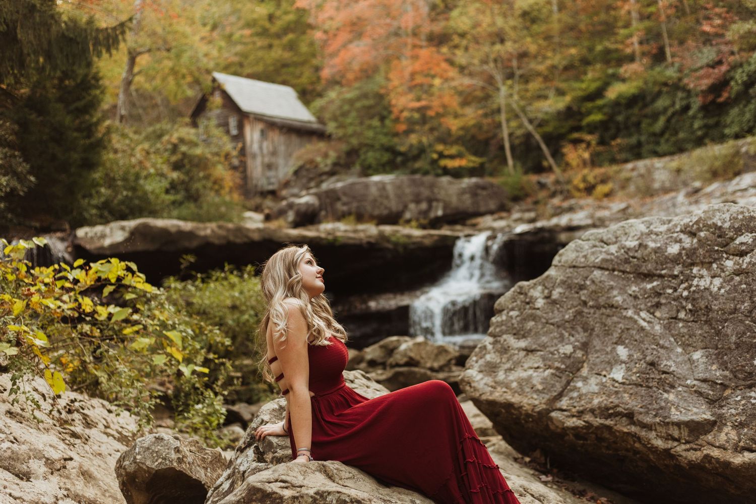 A burgundy gown creates dramatic contrast against rocky cliffs and a cascading waterfall in autumn scenery.