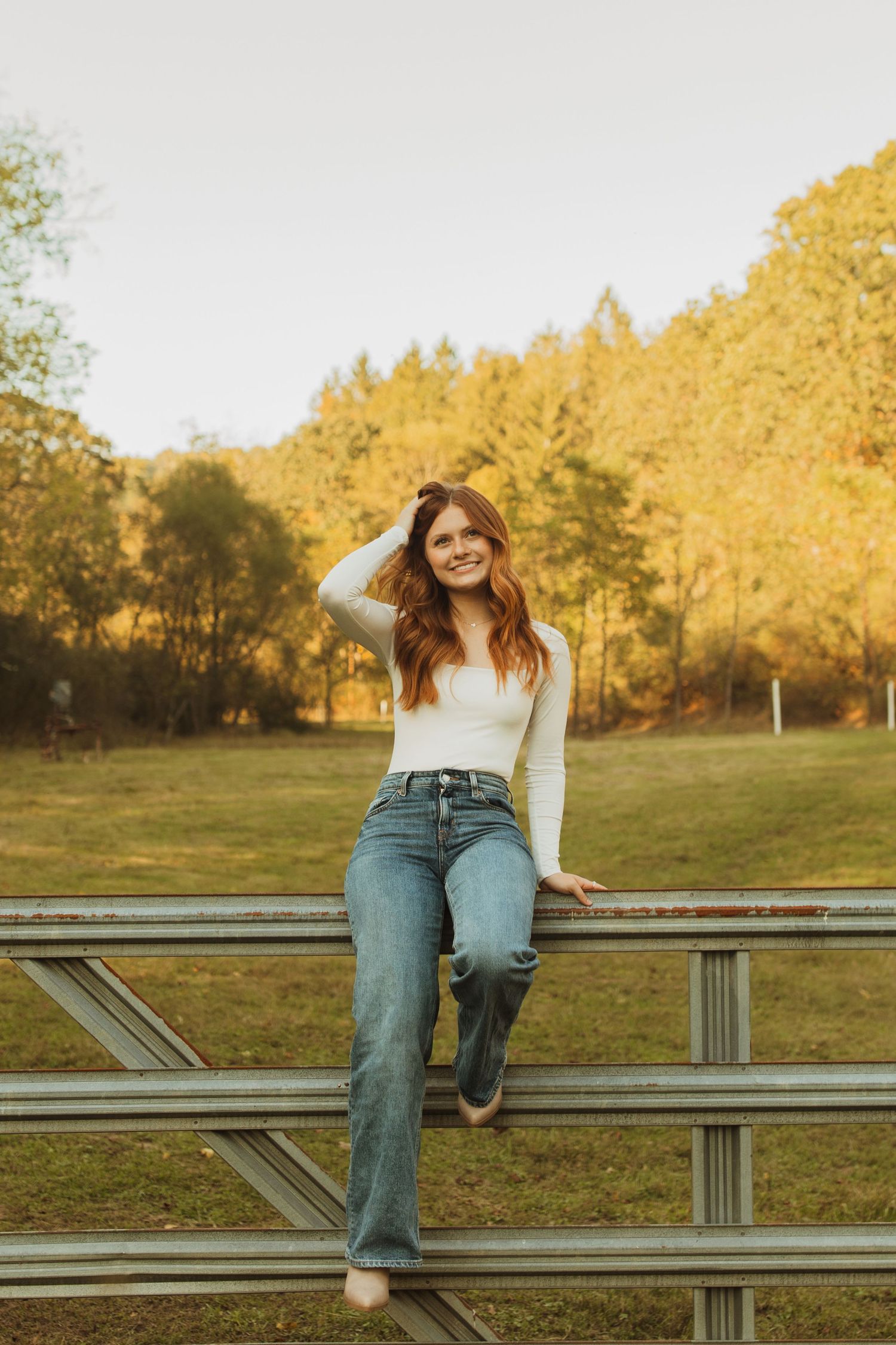 Figure sits casually on wooden fence wearing jeans and white top during golden hour.