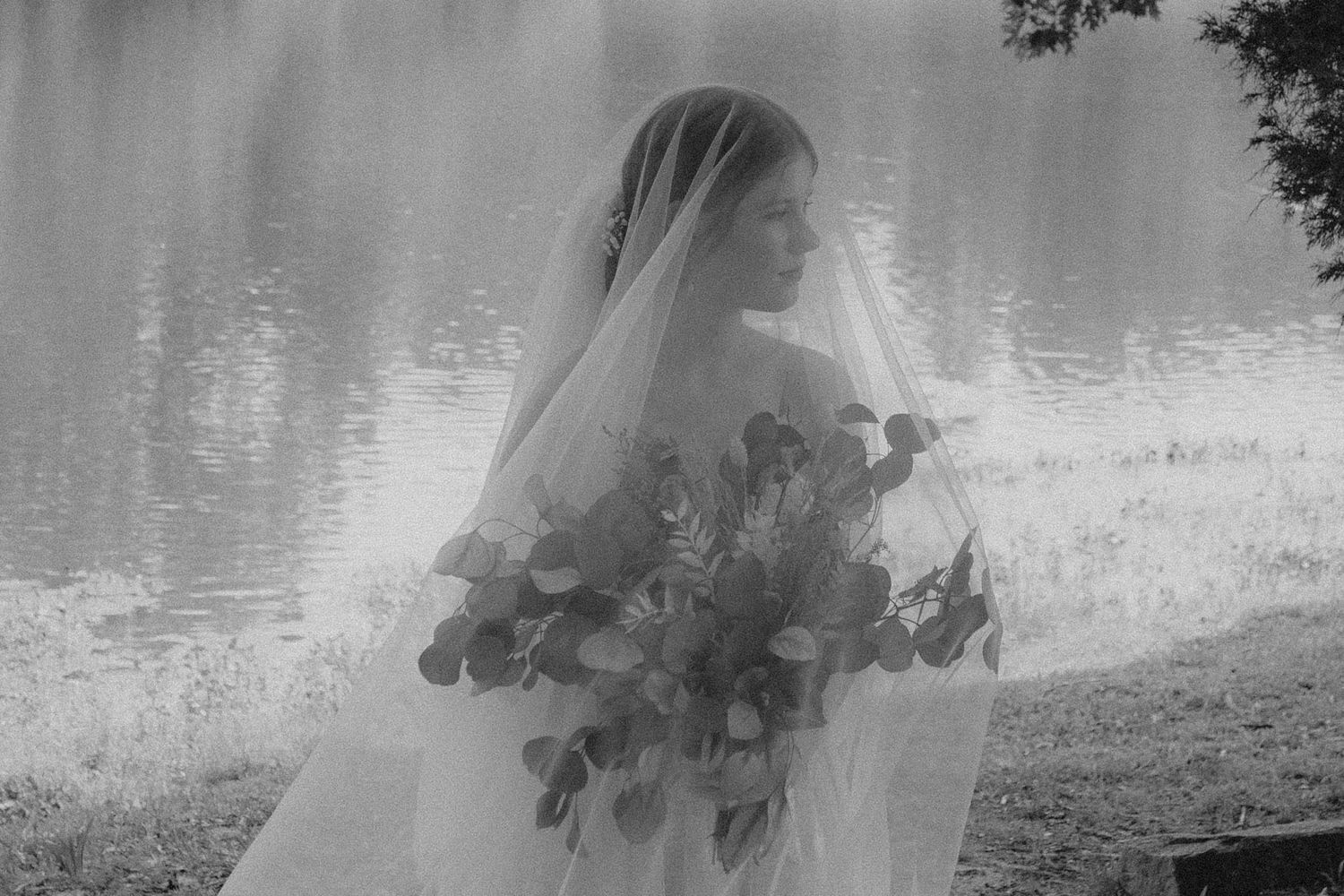 A dreamy black and white bridal portrait showcasing a flowing veil and bouquet beside a serene lake.