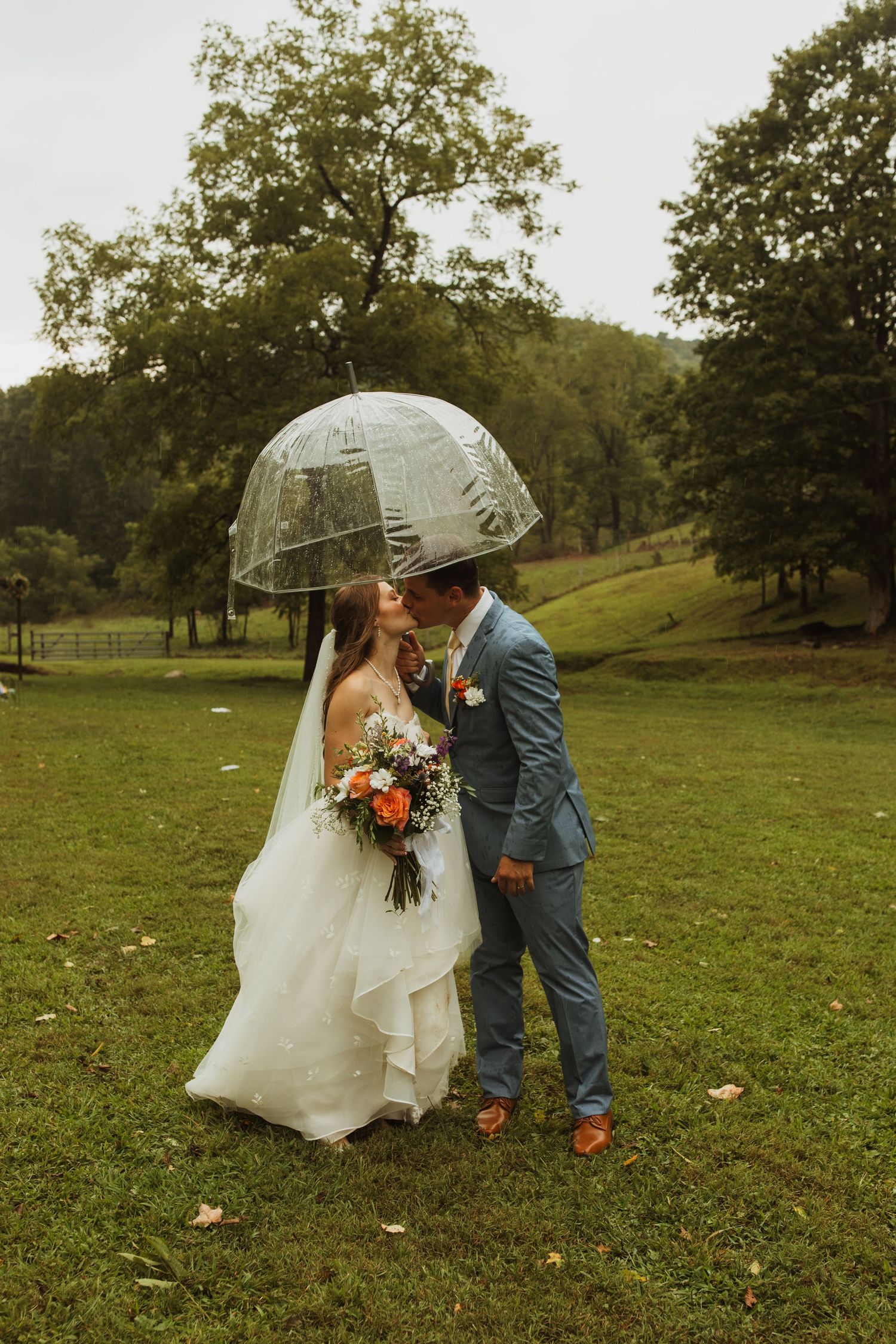 Couple shares kiss under clear umbrella holding vibrant bouquet in rustic field setting.