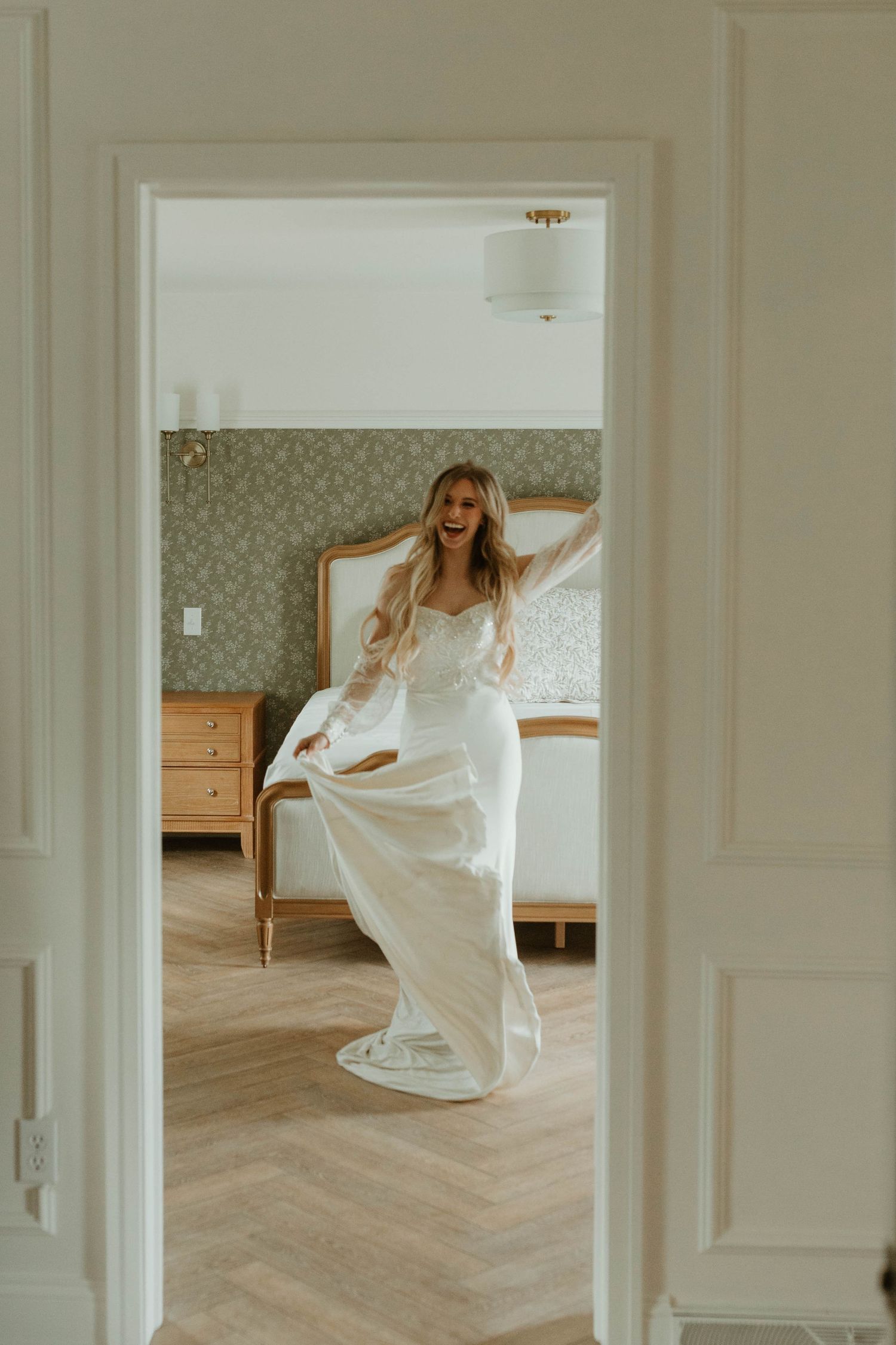 A bride in a flowing white dress twirls playfully in a sunlit bedroom with hardwood floors.