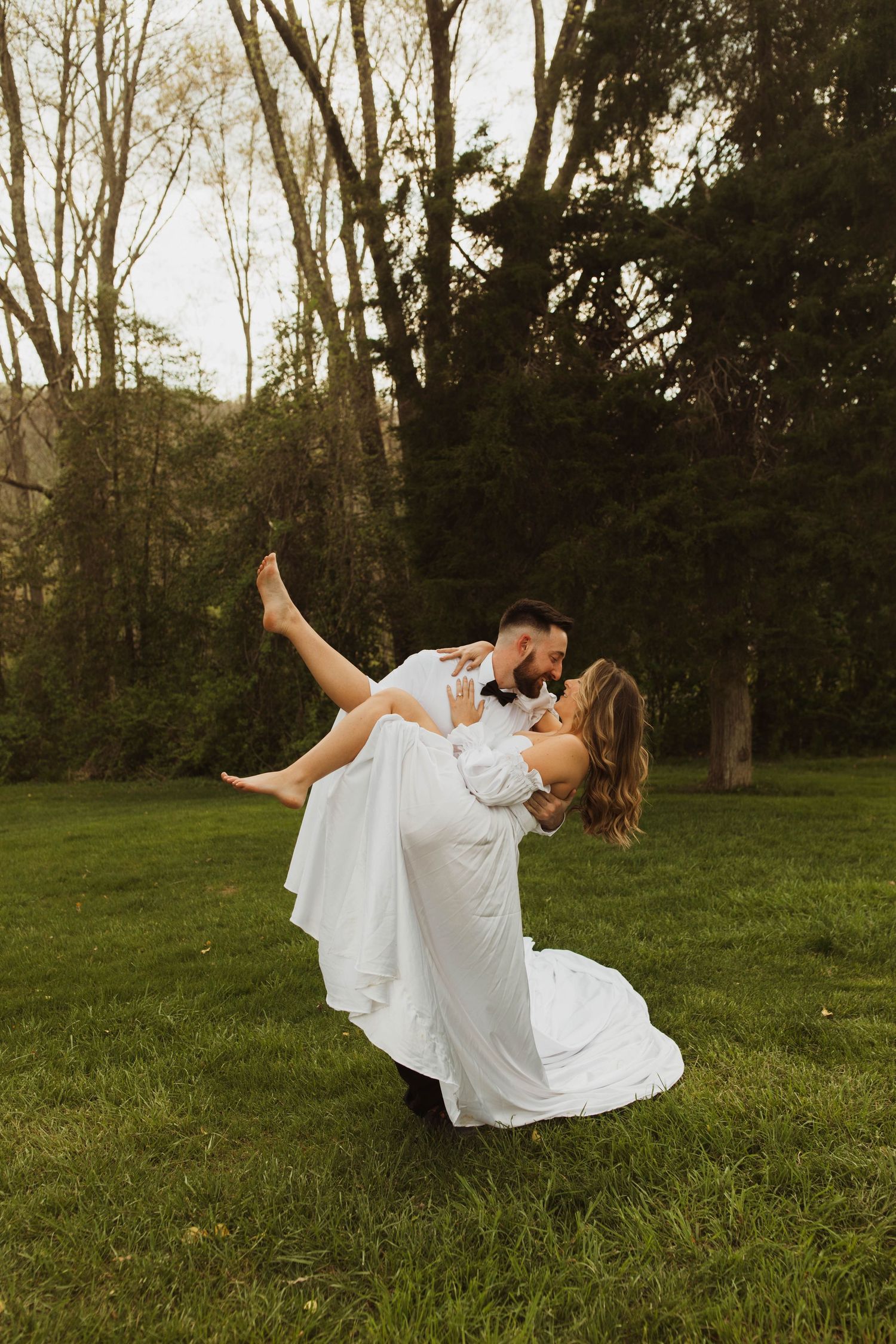 A romantic wedding dance dip pose on a grassy lawn surrounded by trees.