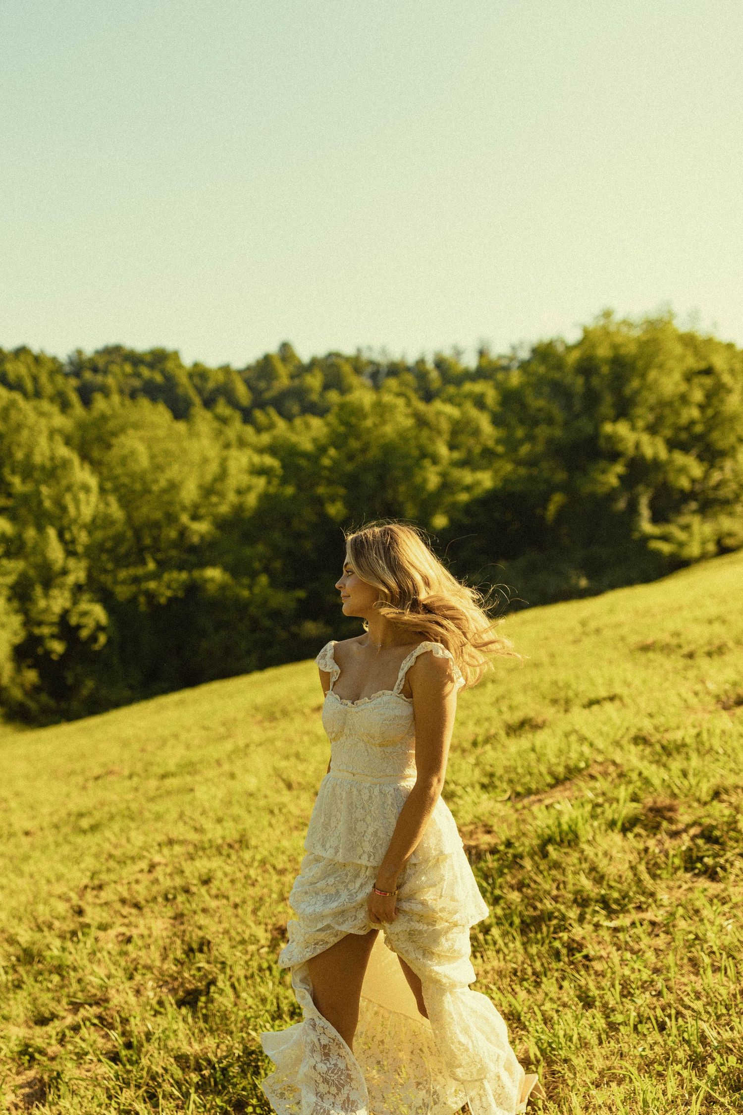 Ethereal portrait of someone in a white dress with wind-swept hair on a sunny hillside.