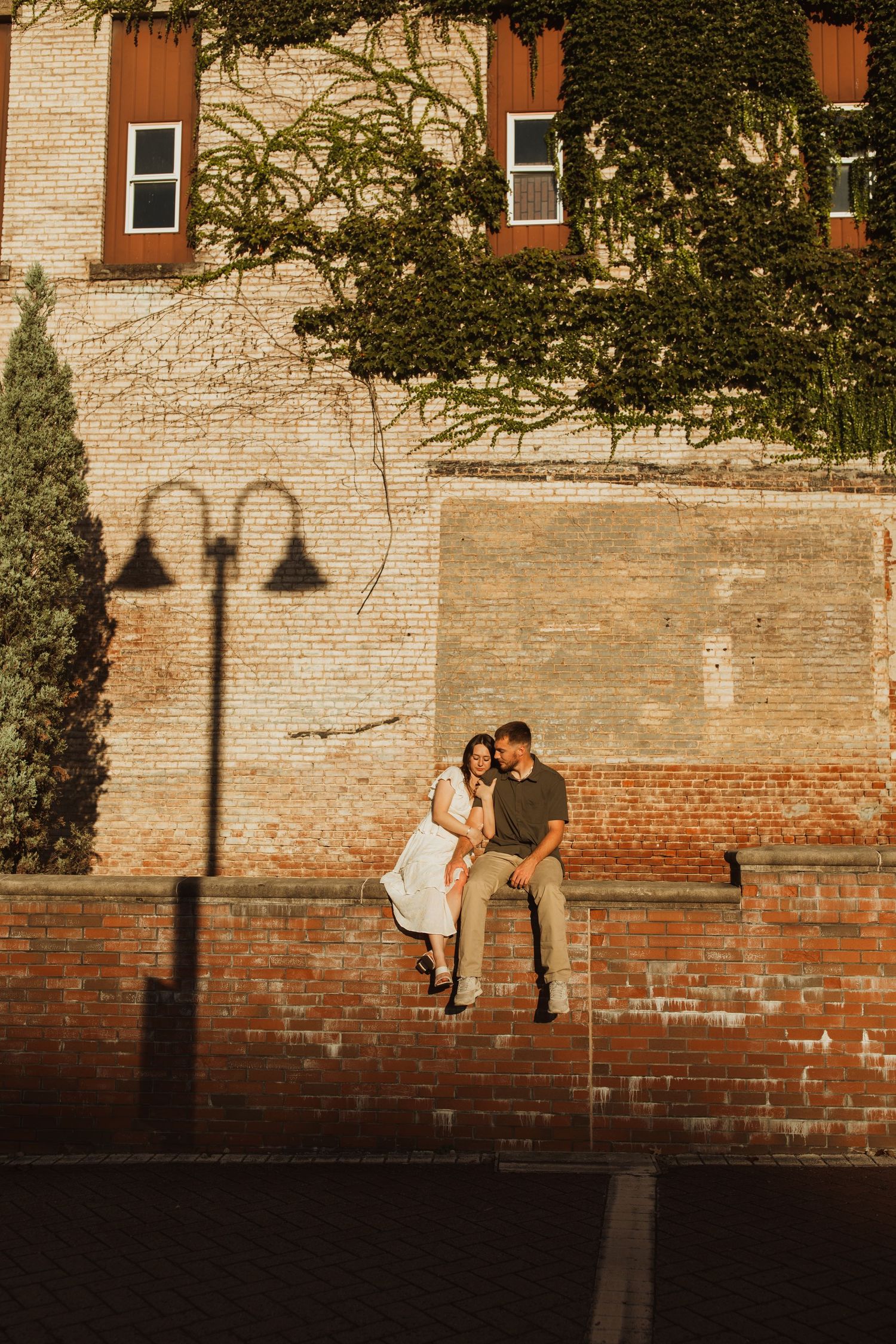 Couple perched on brick ledge against historic building wall at sunset with lamp post shadow.