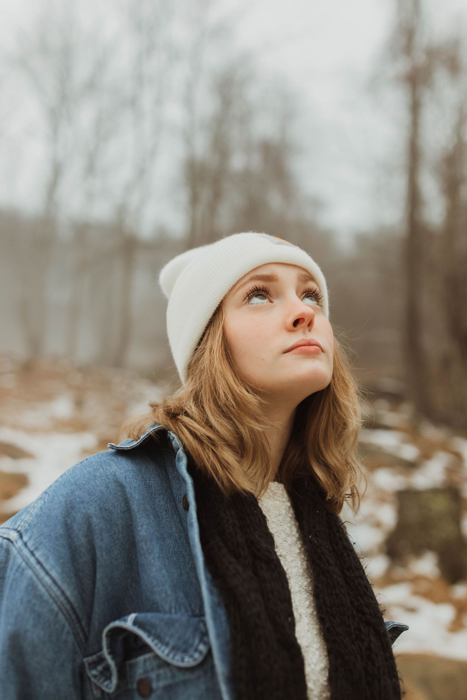 Person in white beanie and denim jacket looking upward against winter forest backdrop.