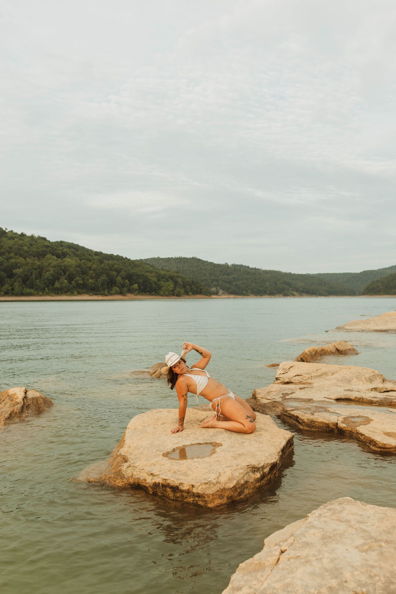 Athletic pose performed on rock formations jutting into a tranquil lake surrounded by hills.