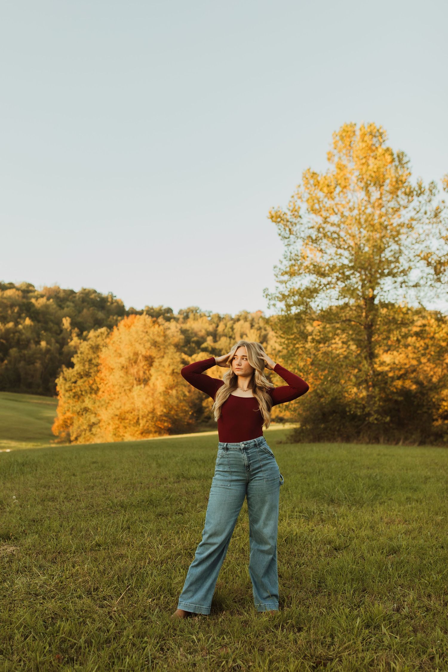 Vintage-inspired pose in bell bottom jeans and burgundy top with golden fall trees in background.