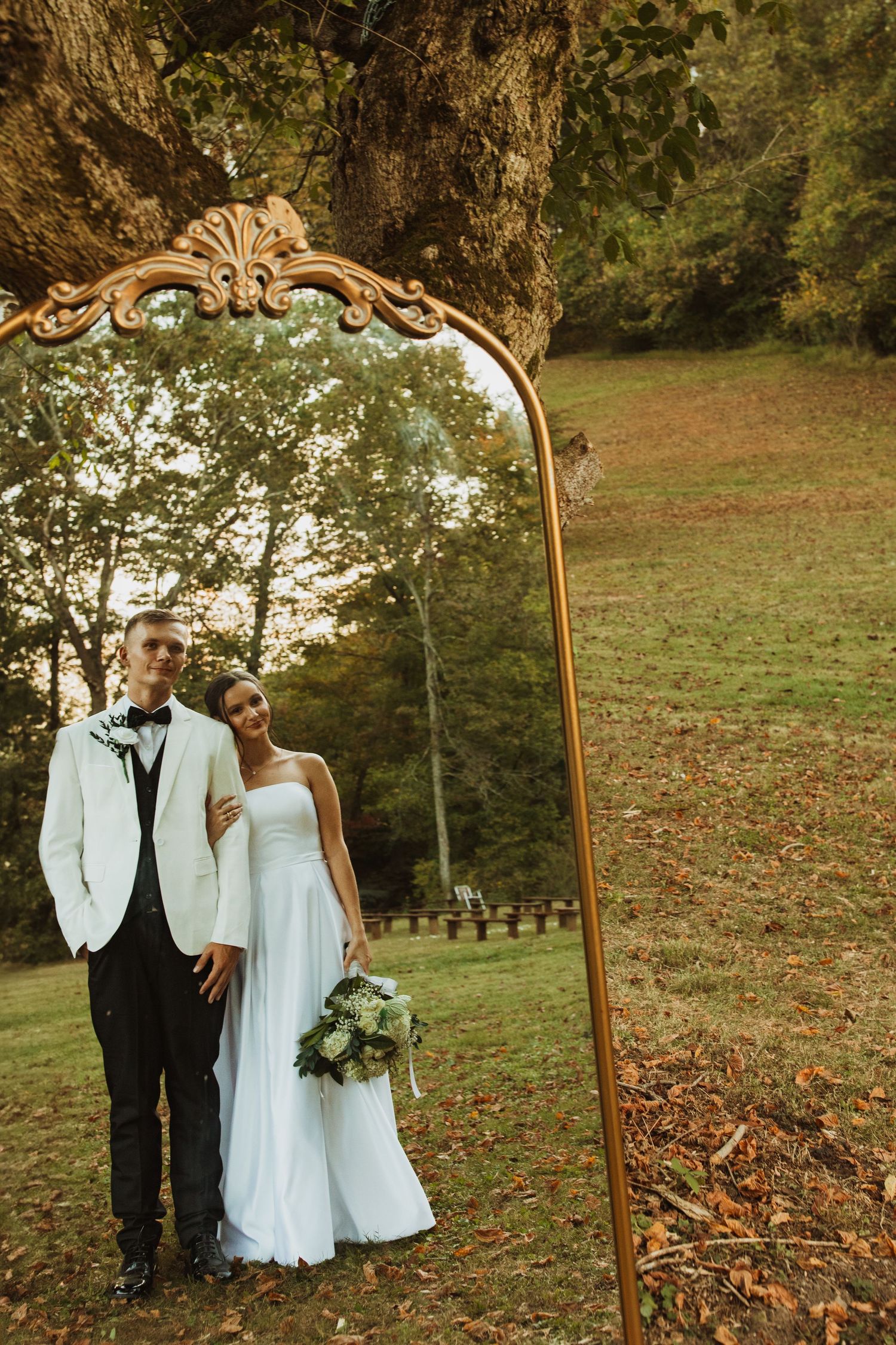A couple poses beside an ornate vintage mirror frame in an autumn outdoor setting.
