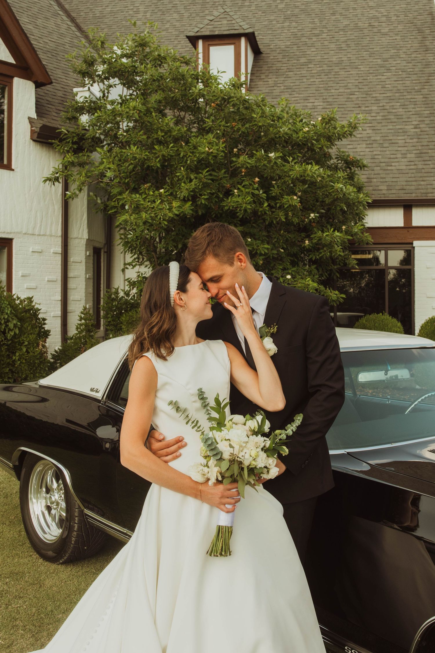 A romantic wedding couple shares an intimate moment in front of a classic black car and Tudor-style house.