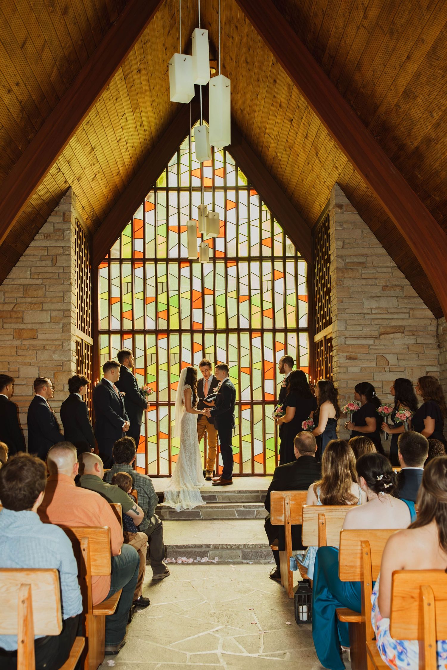 A wedding ceremony in a chapel with a stunning stained glass window backdrop and wooden beamed ceiling.