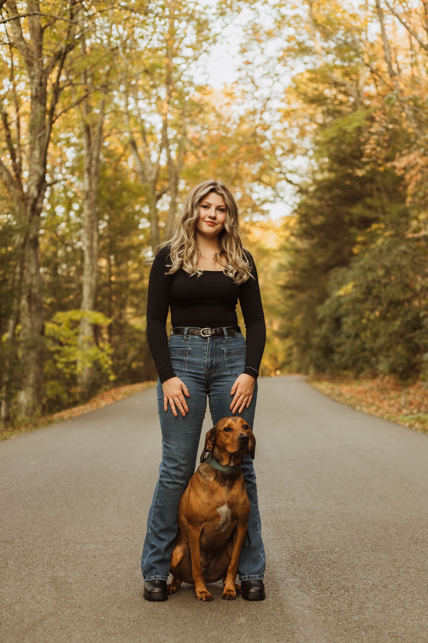 Person in black top and jeans standing with brown dog on autumn forest road.