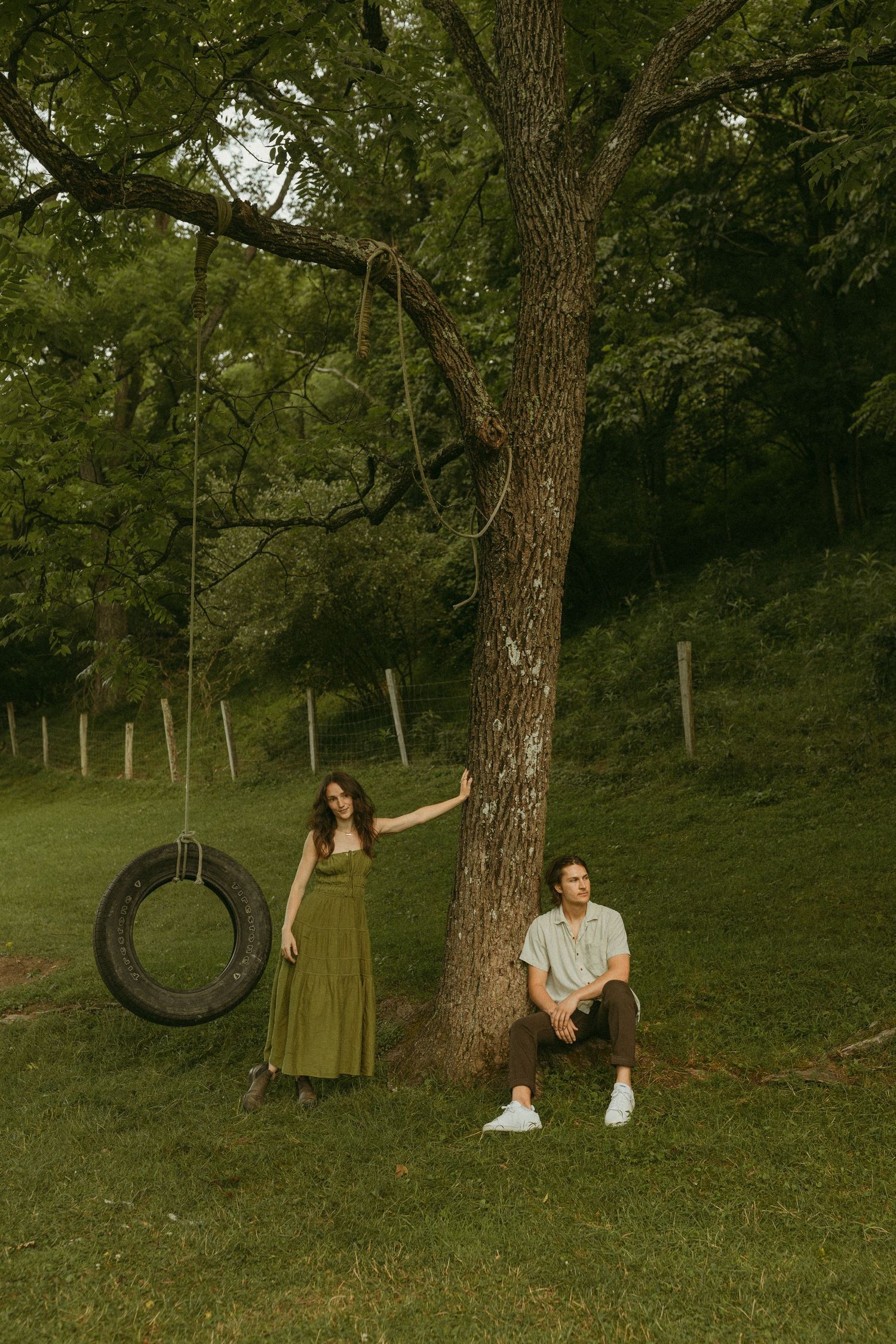 Two people pose artistically by a tire swing hanging from a large tree on a grassy hill.