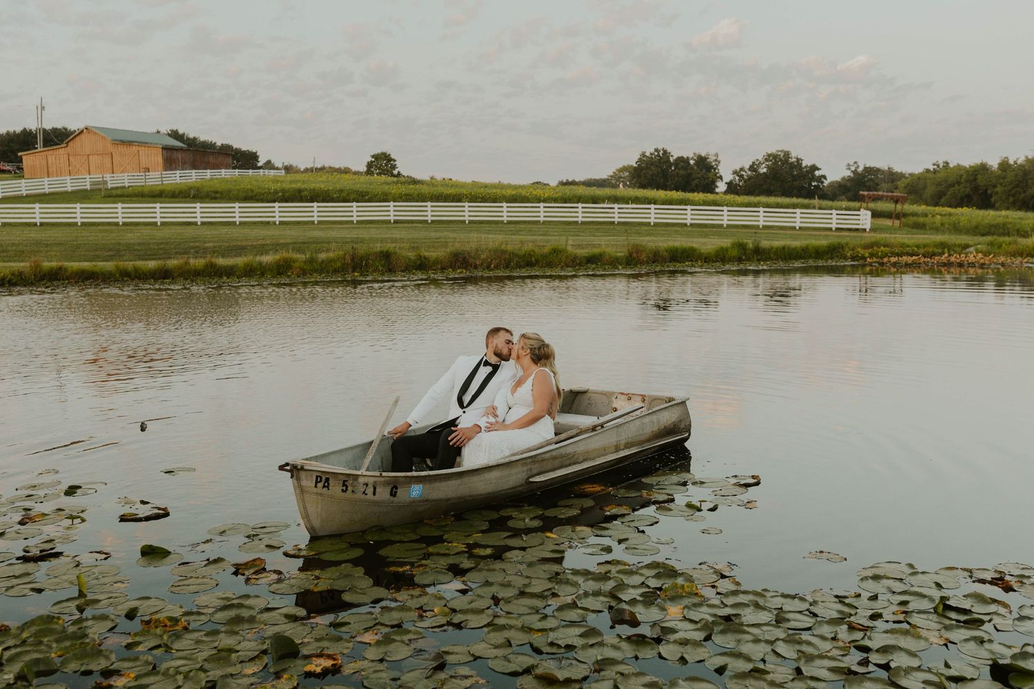 Newlyweds in canoe on pond surrounded by lily pads with white fence in background.