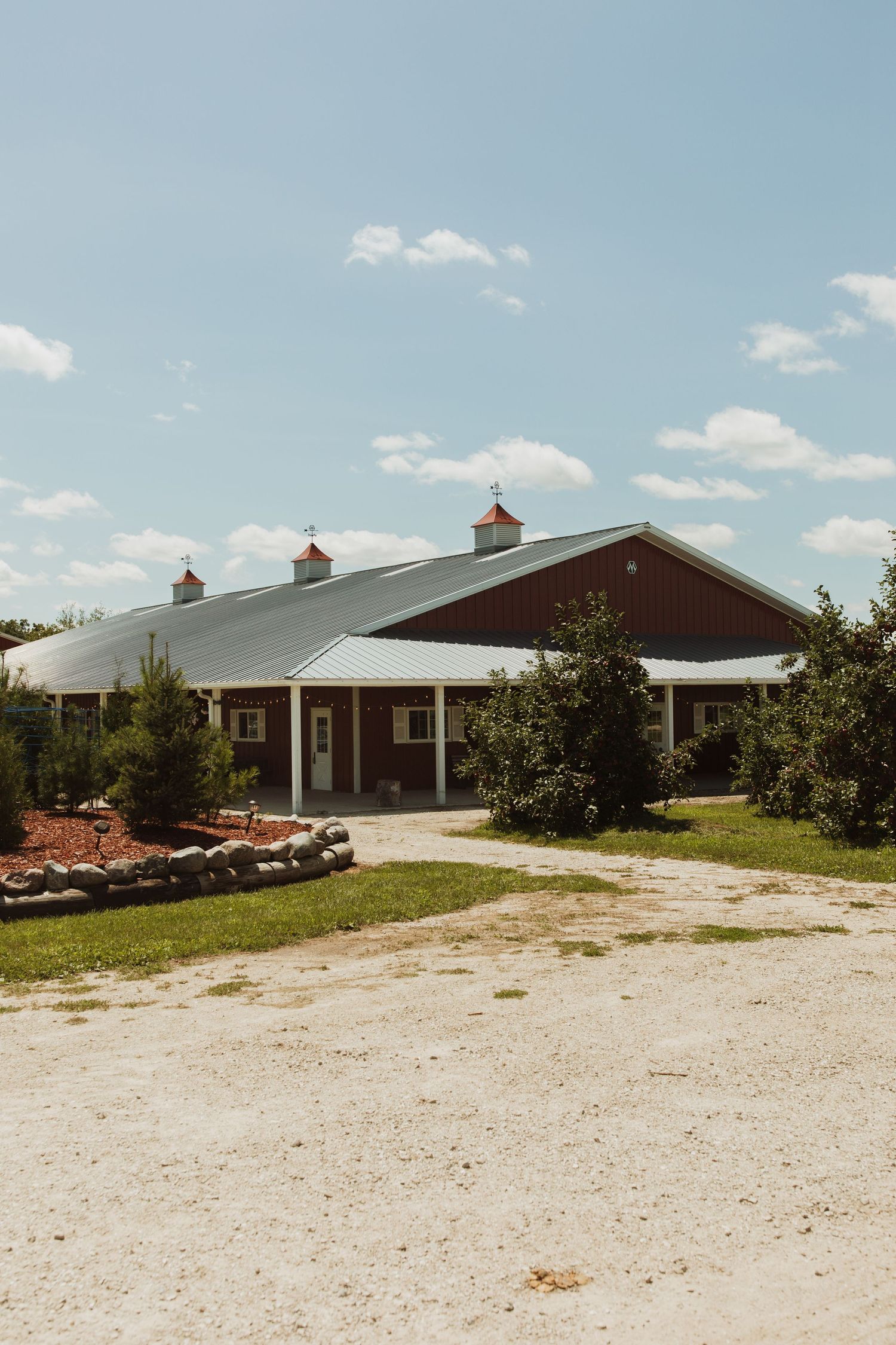 Large red and white barn-style wedding venue with gravel parking area under blue skies.