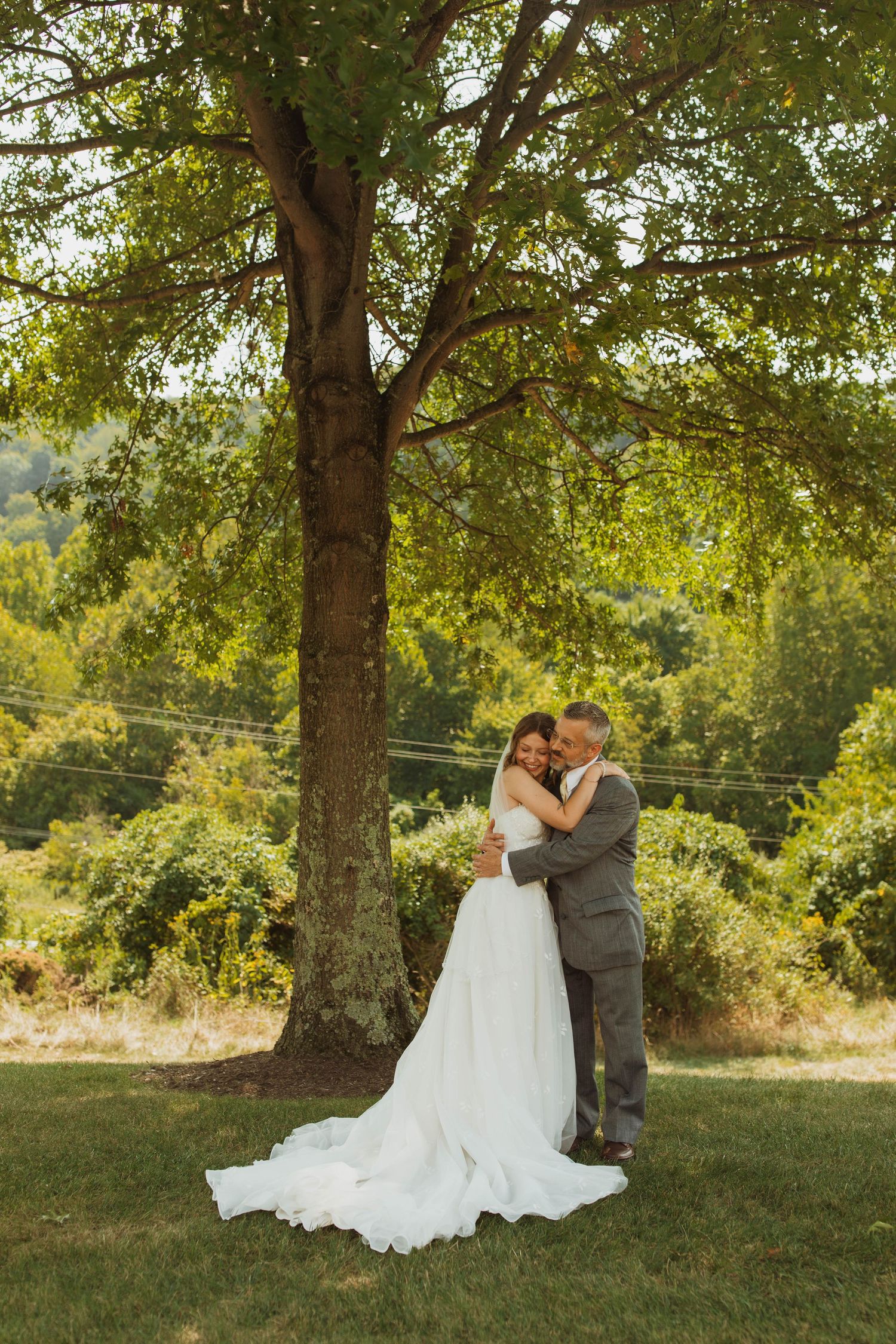 Newlyweds share an intimate moment under a large tree in golden afternoon light.