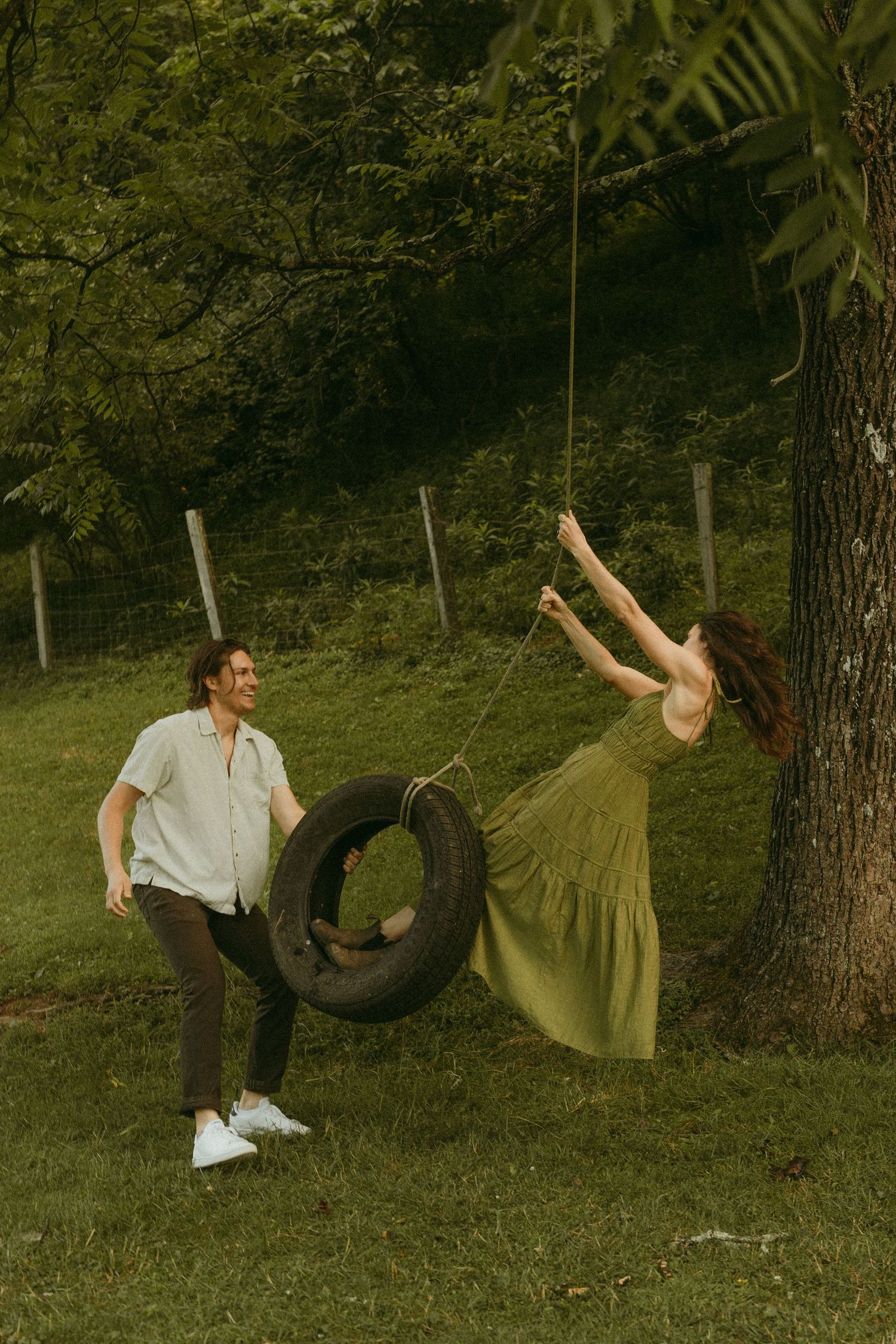 Playful engagement photo with tire swing in a lush green garden setting.