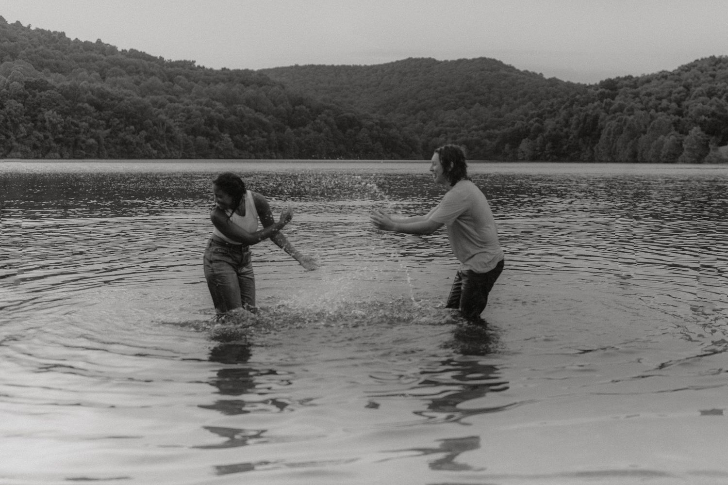 Two figures playing and splashing in a scenic lake surrounded by wooded hills.