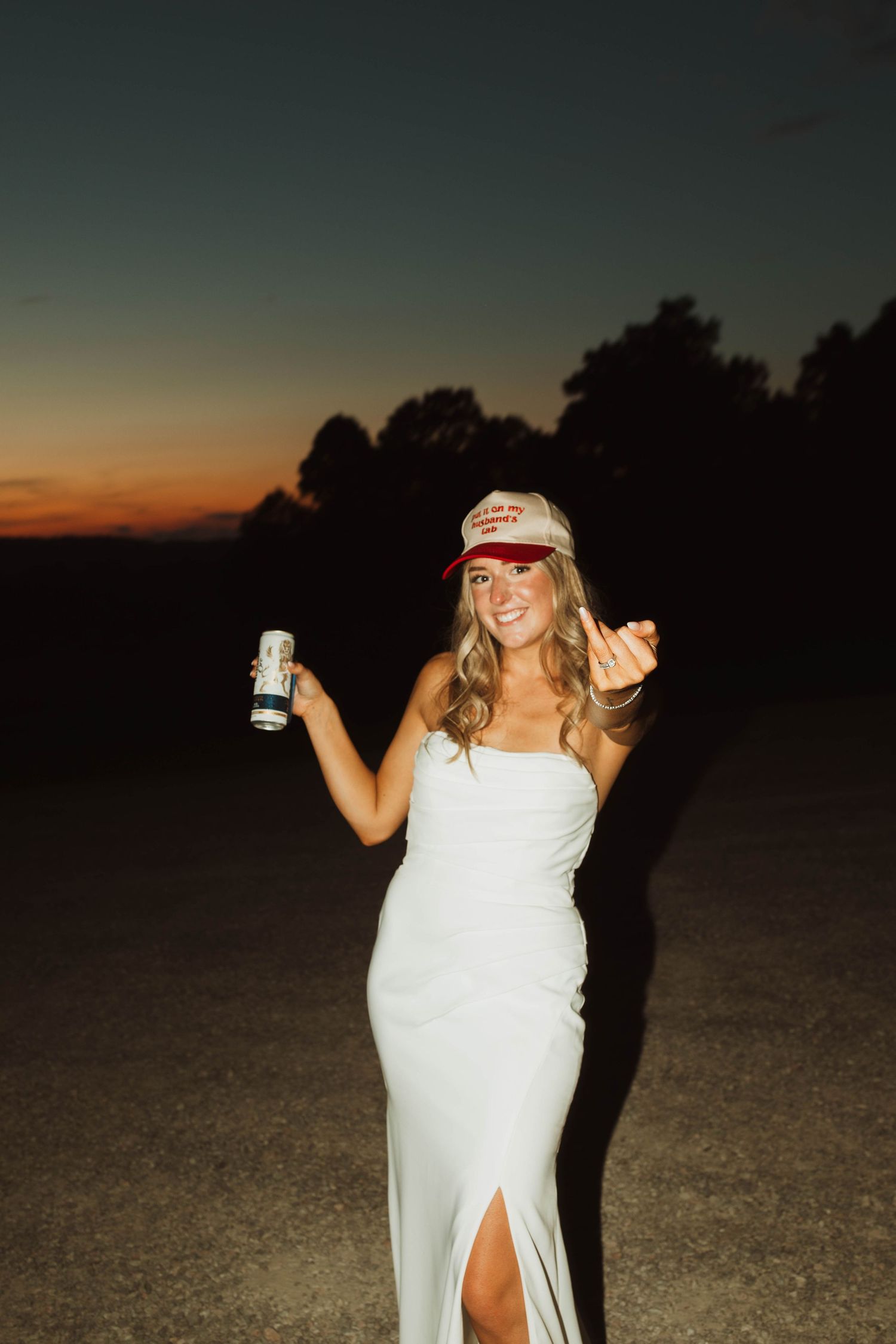 A person in a white dress and baseball cap holds up a drink at sunset.