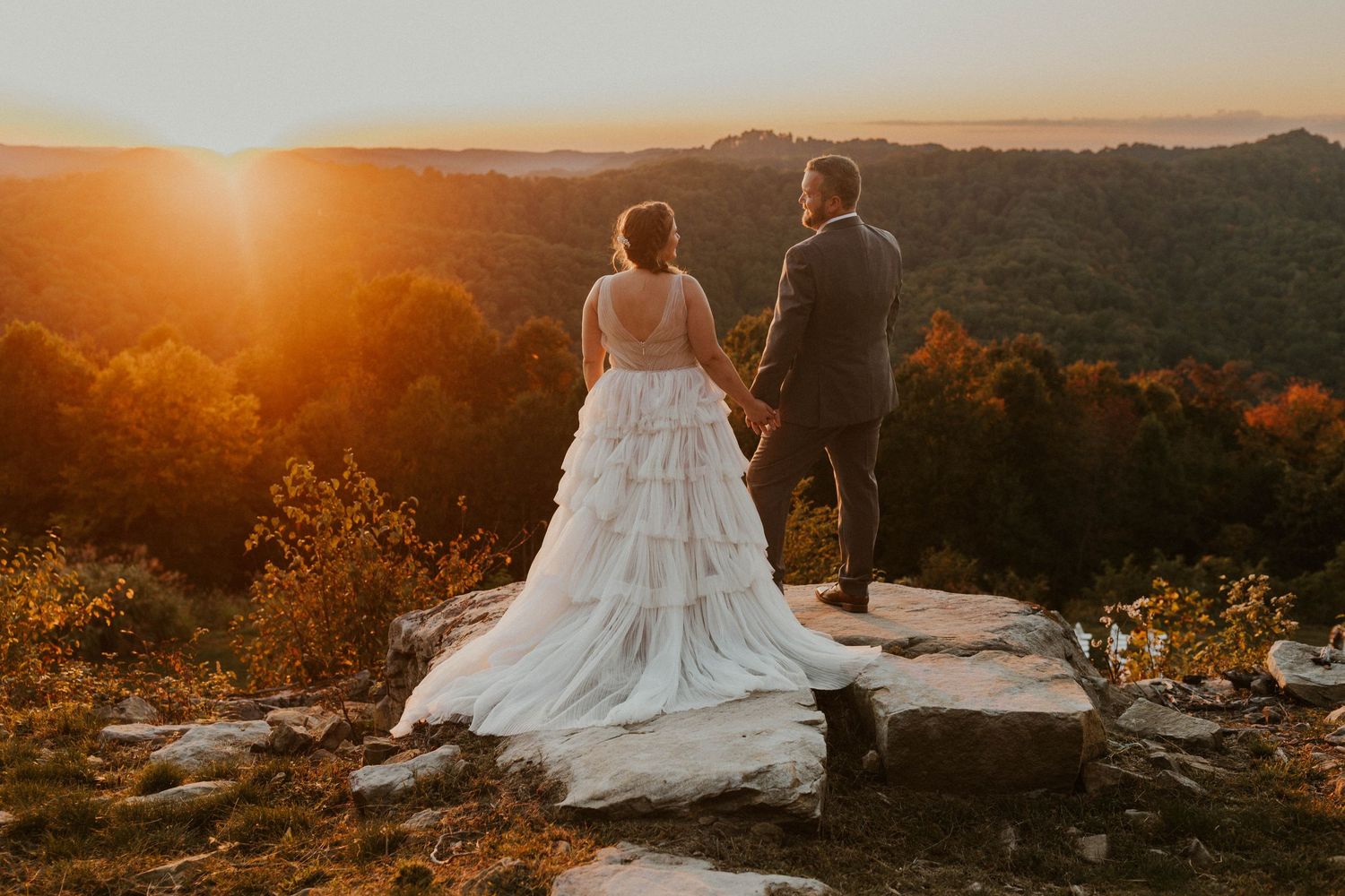 Newlyweds hold hands while admiring mountain vista at sunset.