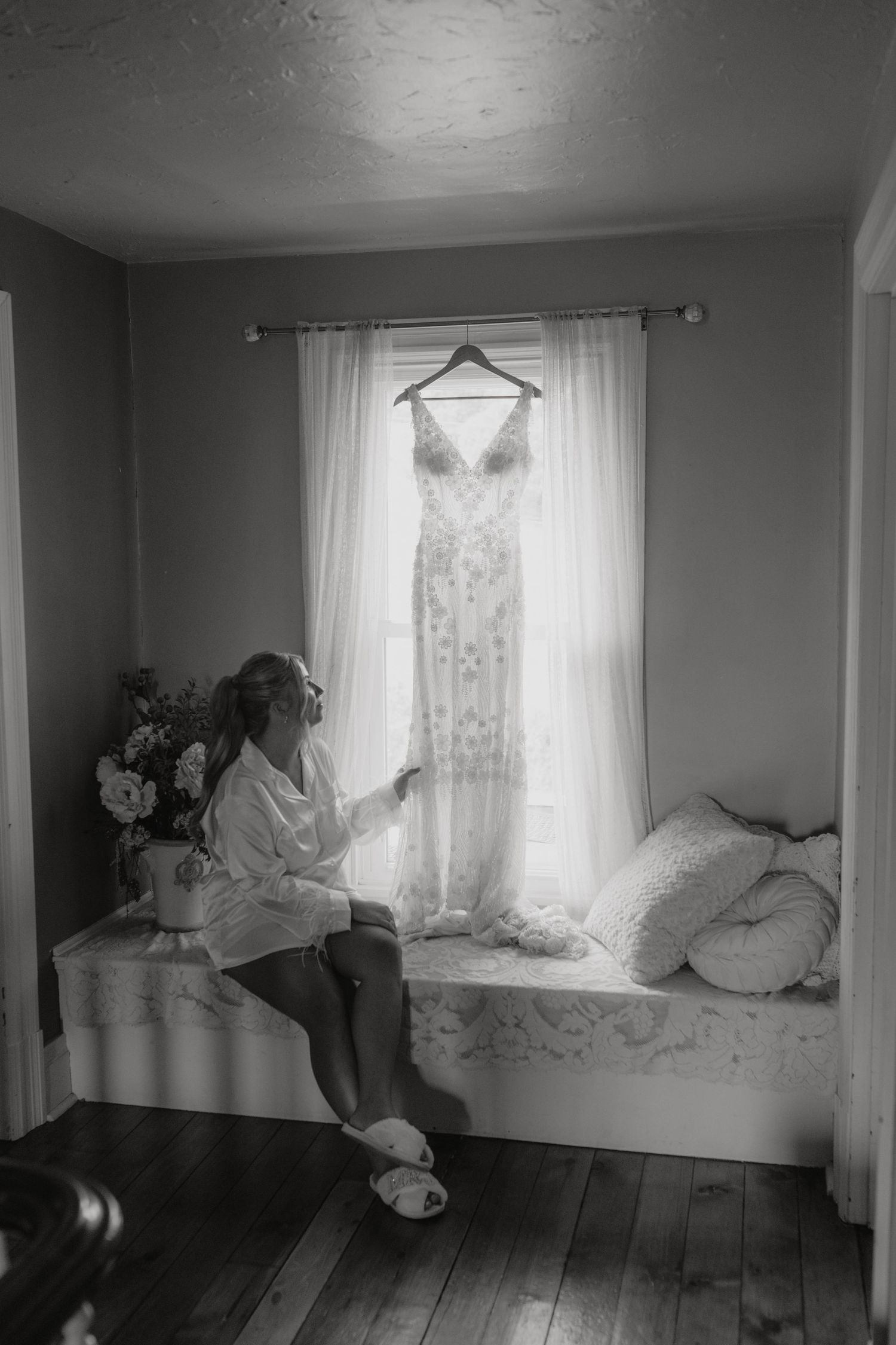 Wedding dress hanging by window while figure sits on bench in morning light.