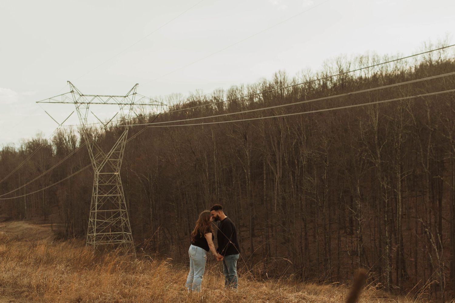 A couple embraces near an electrical transmission tower in a field with bare trees in the background.