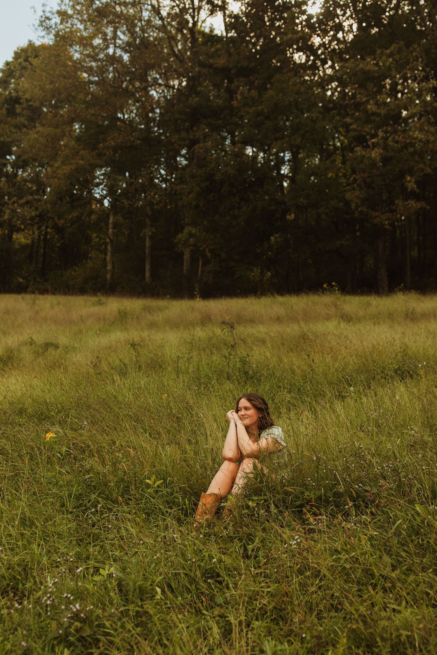 Person in a white dress relaxes in a sunlit meadow with tall grass and trees in the background.