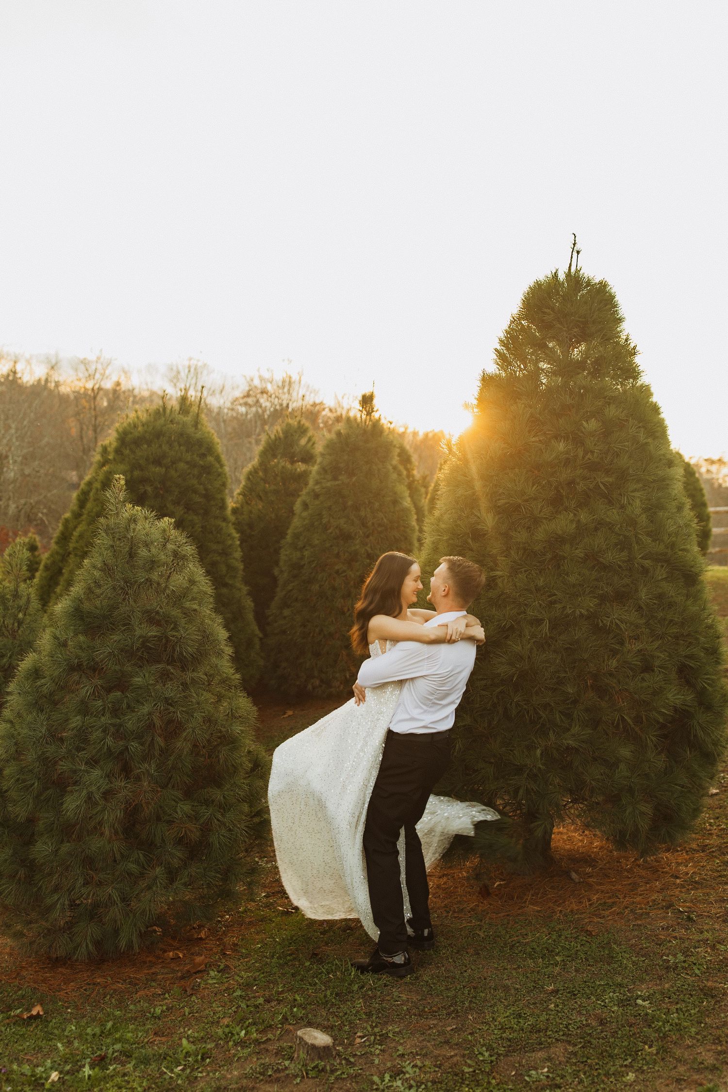 Couple embracing in front of evergreen trees during golden hour sunset at Christmas tree farm.