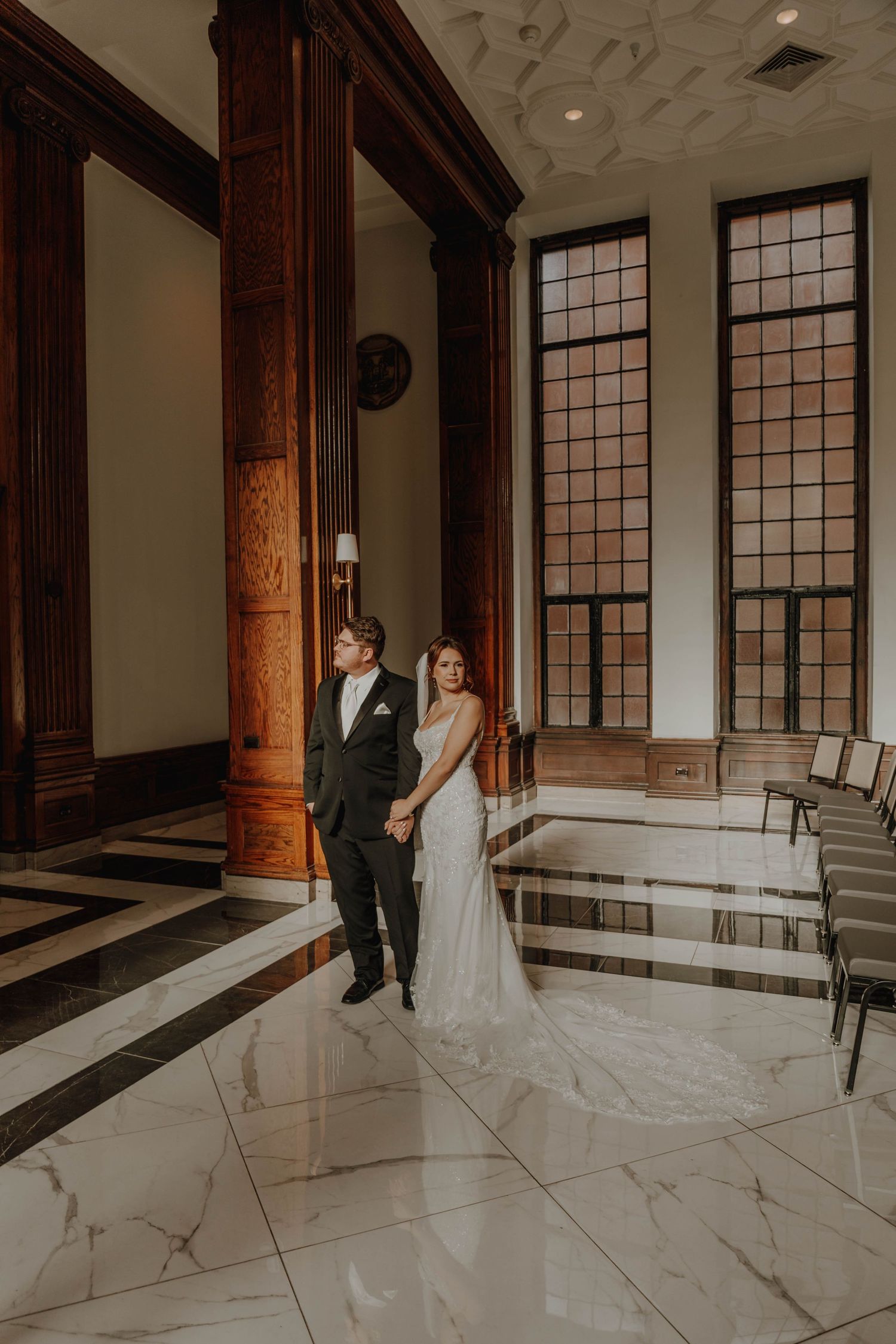 Wedding portrait in a grand hall with marble floors and towering windows.
