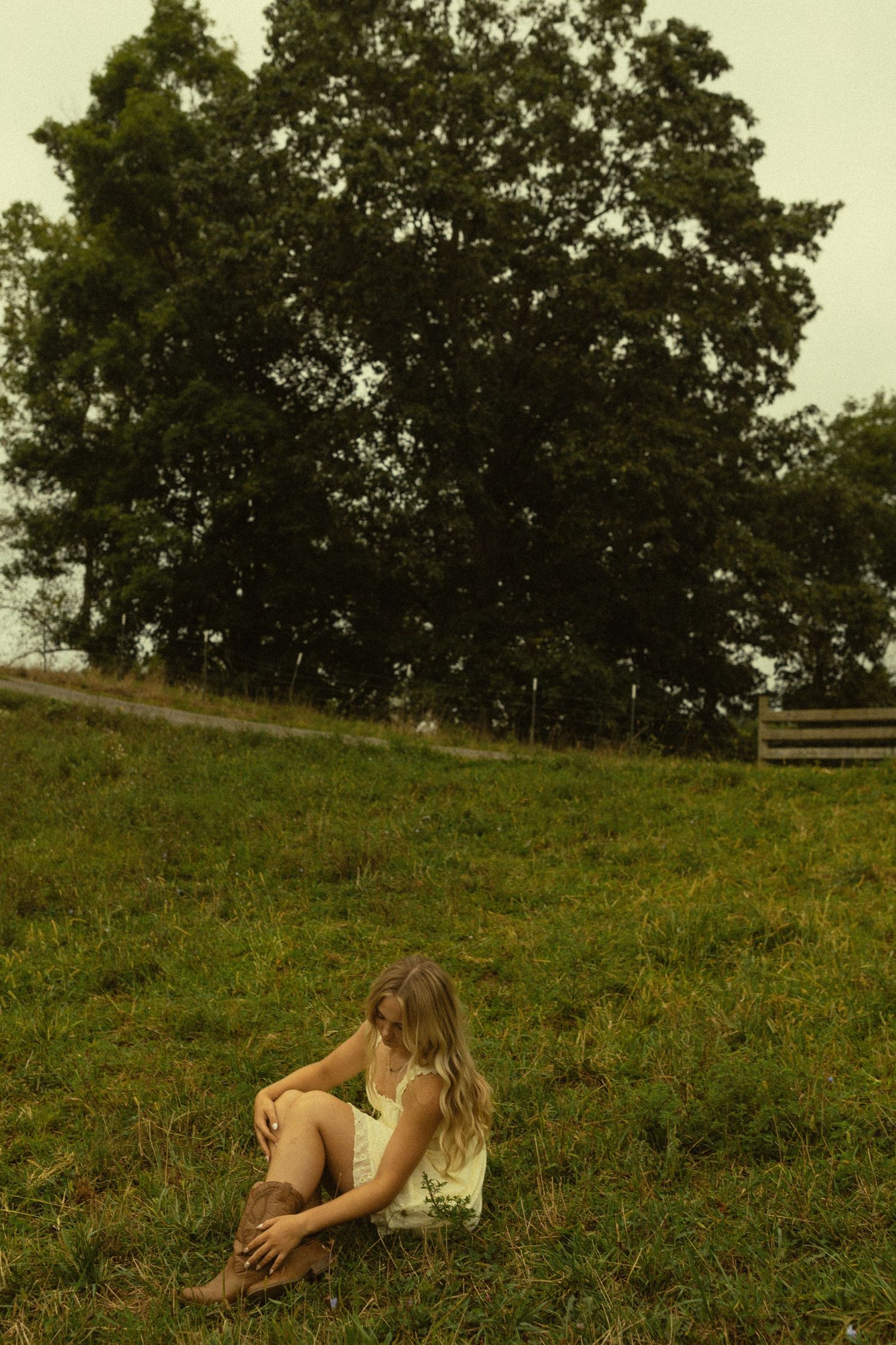 Figure in white dress sits thoughtfully on grassy hillside near wooden fence.