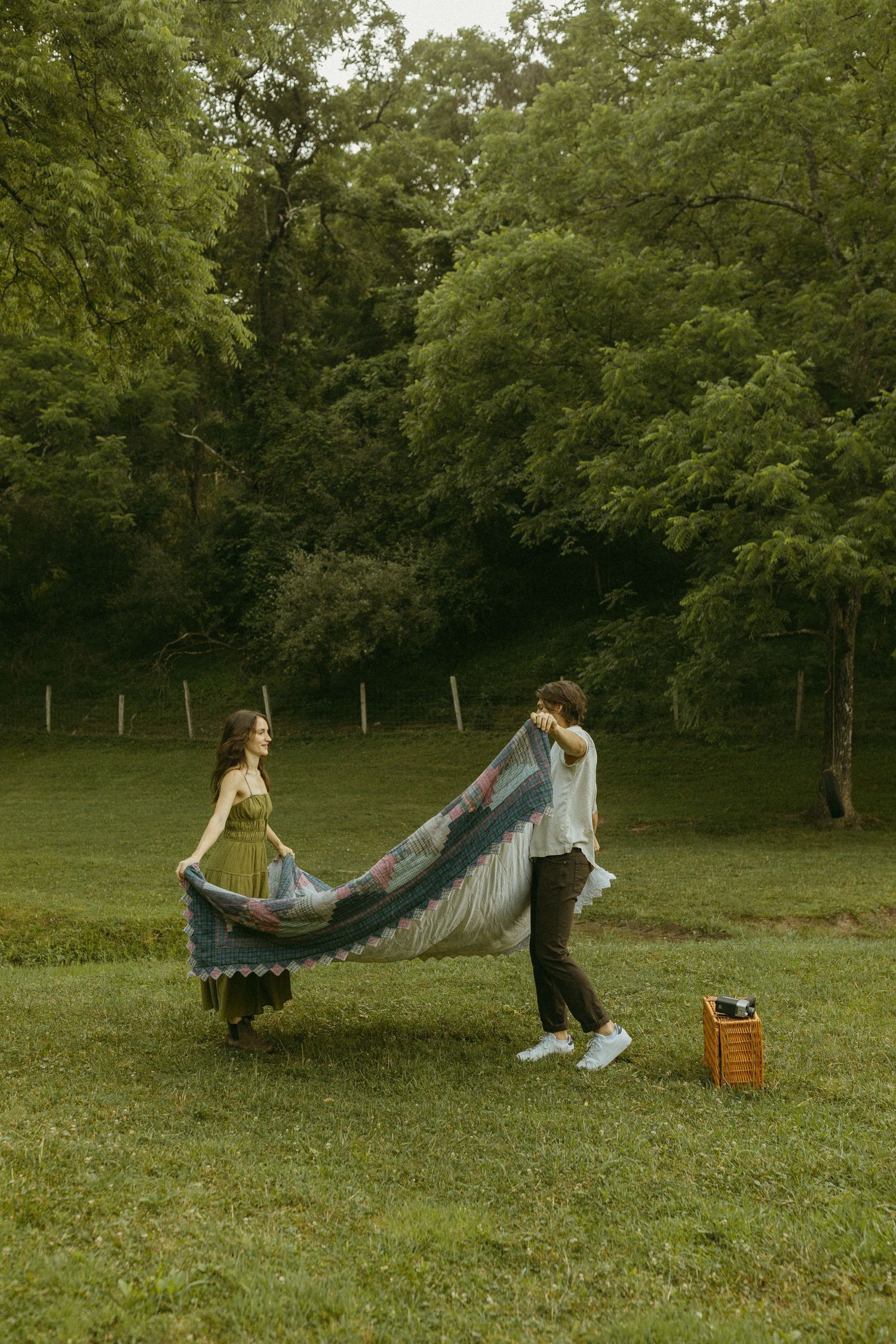 A couple in casual attire shakes out a blue blanket together on a grassy field surrounded by trees.