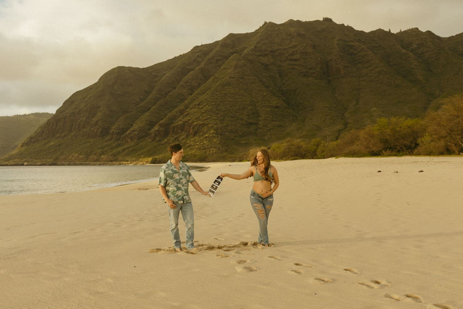 Couple holds hands while walking on sandy beach with dramatic mountain backdrop.